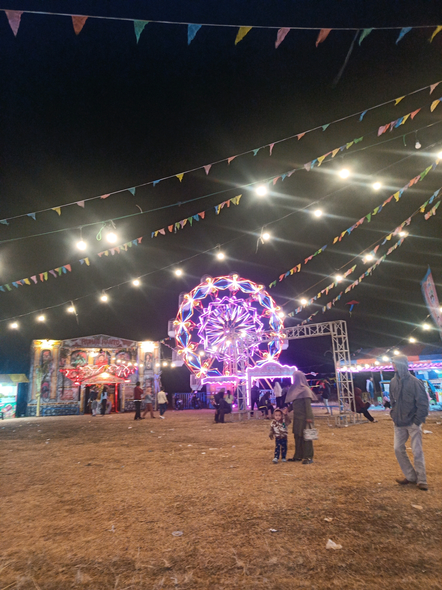 The image depicts a nighttime scene at what appears to be a local fair or carnival in Tanjungsari, Indonesia. Dominating the center of the frame is a brightly lit Ferris wheel, adorned with colorful LED lights, casting a vibrant glow on the surrounding area. In the background, a building with an ornate facade, possibly an attraction or stall, is illuminated by strings of lights and festive banners are strung across the dark sky. Several people are visible, scattered across the dusty ground, some walking, others standing near the attractions. In the foreground, a person in a hooded jacket stands to the right, while to the left, an adult wearing a hijab is accompanied by a small child. The ground is covered in dry grass and scattered debris. The overall atmosphere suggests a lively community gathering under the night sky, illuminated by the festive lights of the carnival rides and decorations.