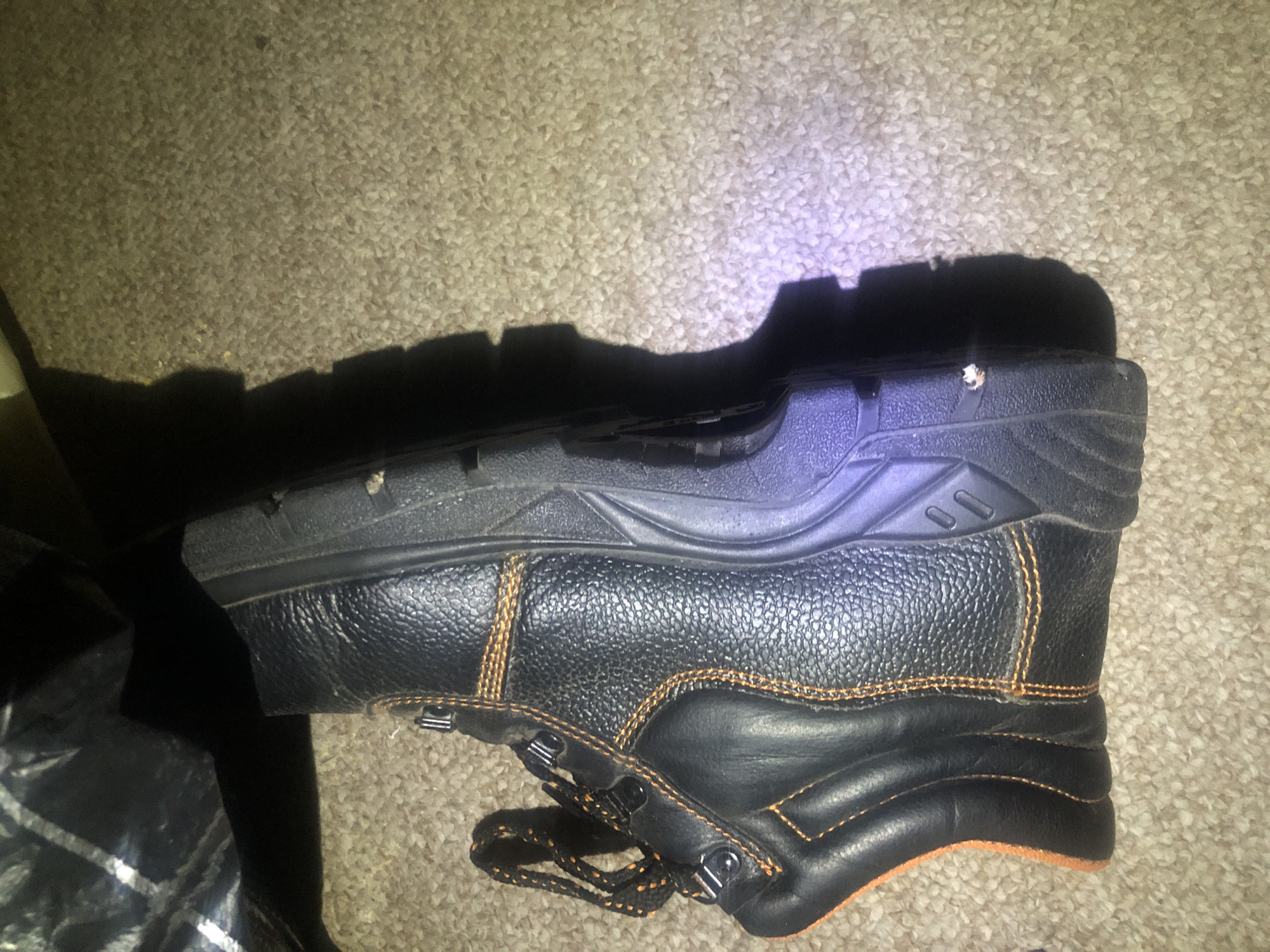 A black leather work boot with orange stitching and an orange accent on the heel is lying on its side on a beige carpeted floor. The boot has black laces with orange threading and silver eyelets. The sole is black and appears to be made of rubber. A dark, possibly plastic, object is partially visible in the upper left corner of the image, casting a shadow over the boot. The background is a carpeted floor with visible fibers and a slight texture. There is a purple light reflection on the sole of the boot. There are no people present in the image. The location in Jalingo, Nigeria, is unknown.