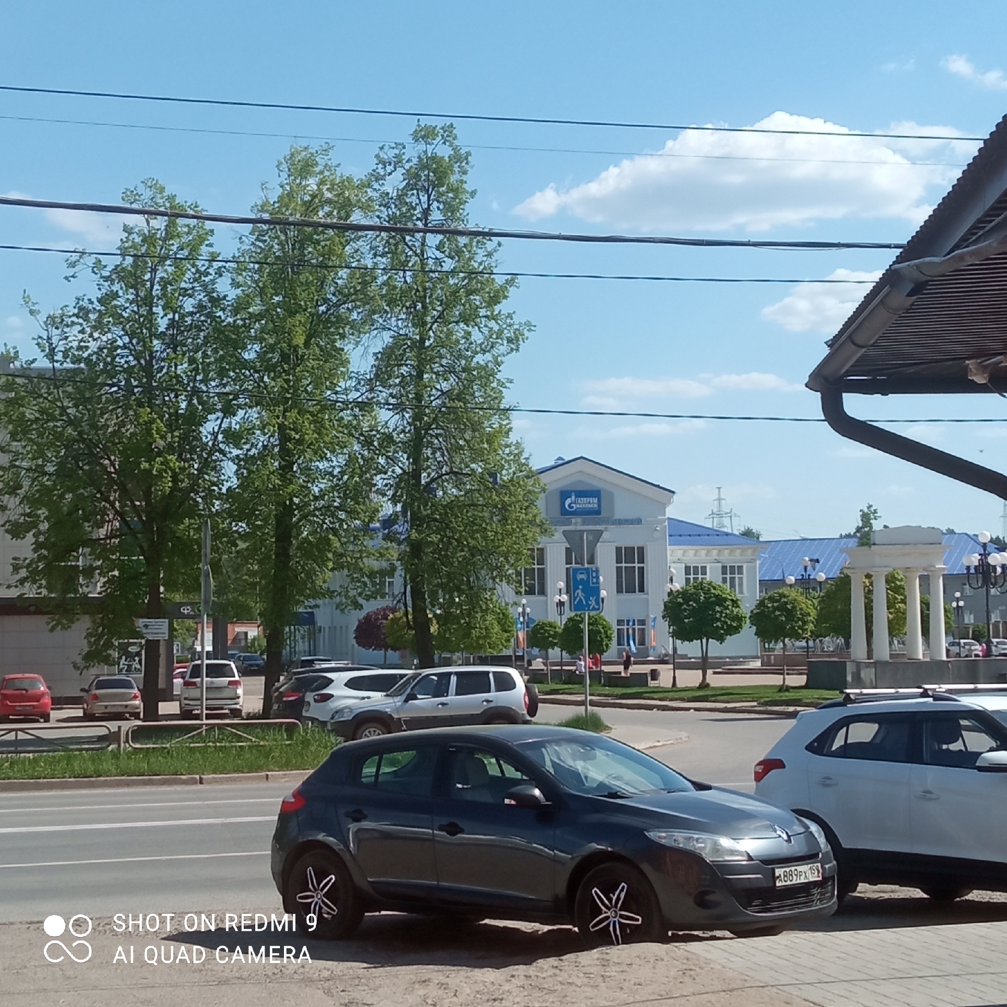The image depicts a street scene in Chaikovsky, Russia, featuring a dark gray Renault Megane parked on the side of the road. A white Hyundai Creta is partially visible beside it. In the background, across the street, is the Gazprom Mezhregiongaz Perm branch building, easily identified by its blue roof and the Gazprom logo. A white colonnade structure, possibly part of a fountain or monument, is visible to the right of the Gazprom building. Trees line the street, and various other cars are parked in the distance. The sky is blue with scattered clouds. Power lines are visible above the trees. The license plate of the Renault Megane is visible: "A889PX159".