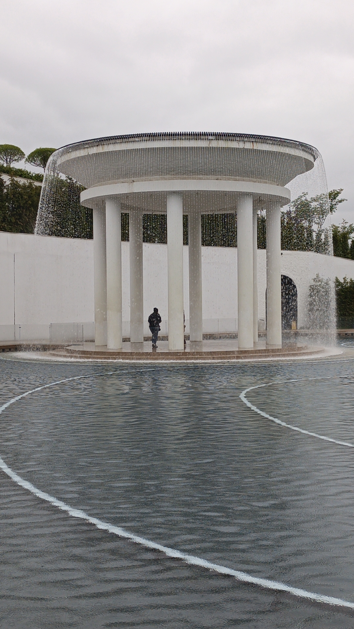 The image shows a circular pavilion with a water feature in what appears to be a public space, possibly in Krasnodar, Russia. The pavilion is a circular structure with white columns supporting a flat roof from which water cascades down, creating a curtain effect. A person is standing under the pavilion, near the edge of the raised platform. The pavilion is surrounded by a shallow pool of water with concentric white lines. A white wall and greenery are visible in the background. The sky is overcast, suggesting a cloudy day.