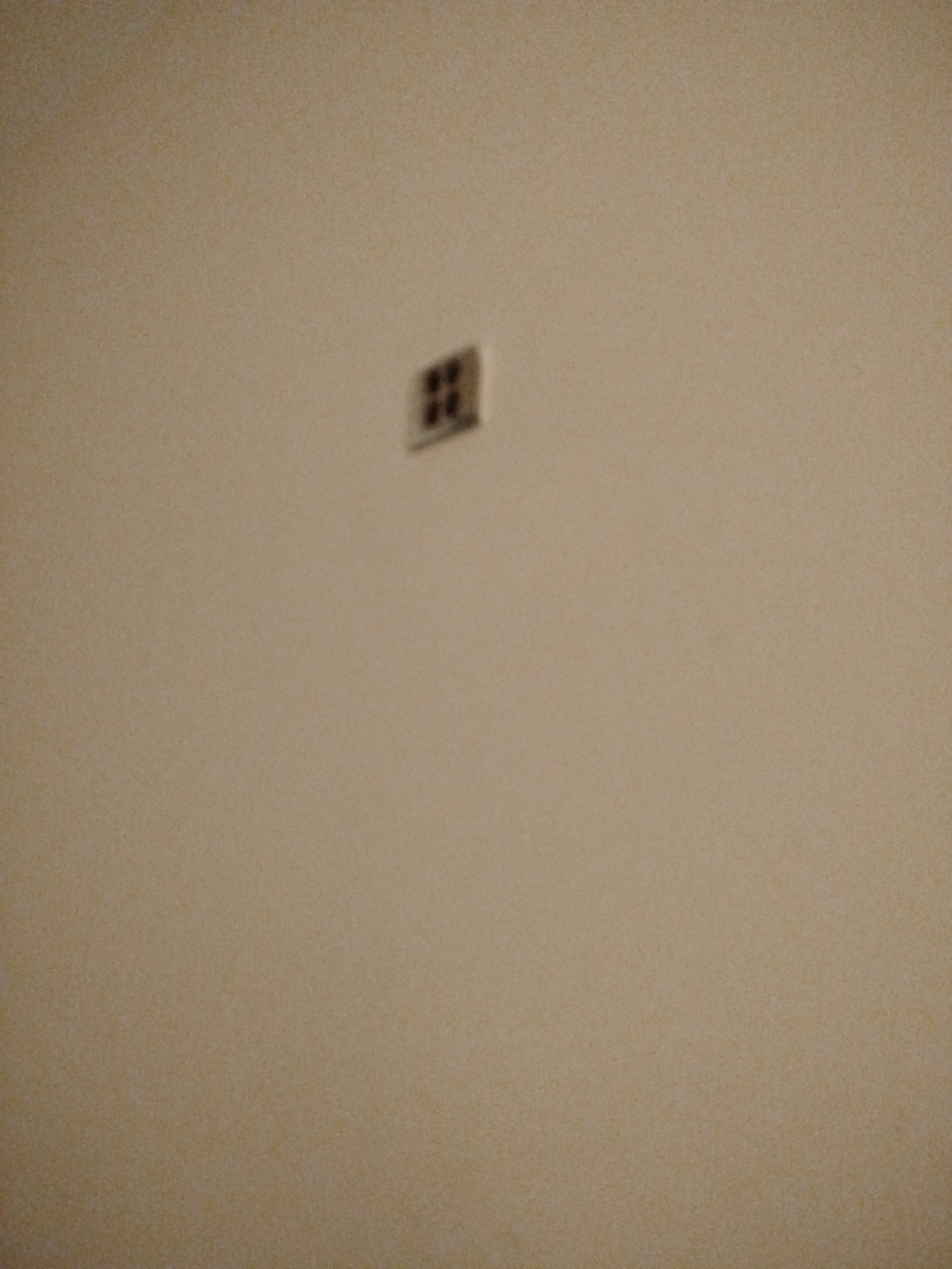 A very small, dark, square object with a pattern of four smaller squares within it is attached to a light tan, textured wall. The object is centrally located in the upper half of the frame. The wall appears to be indoors.