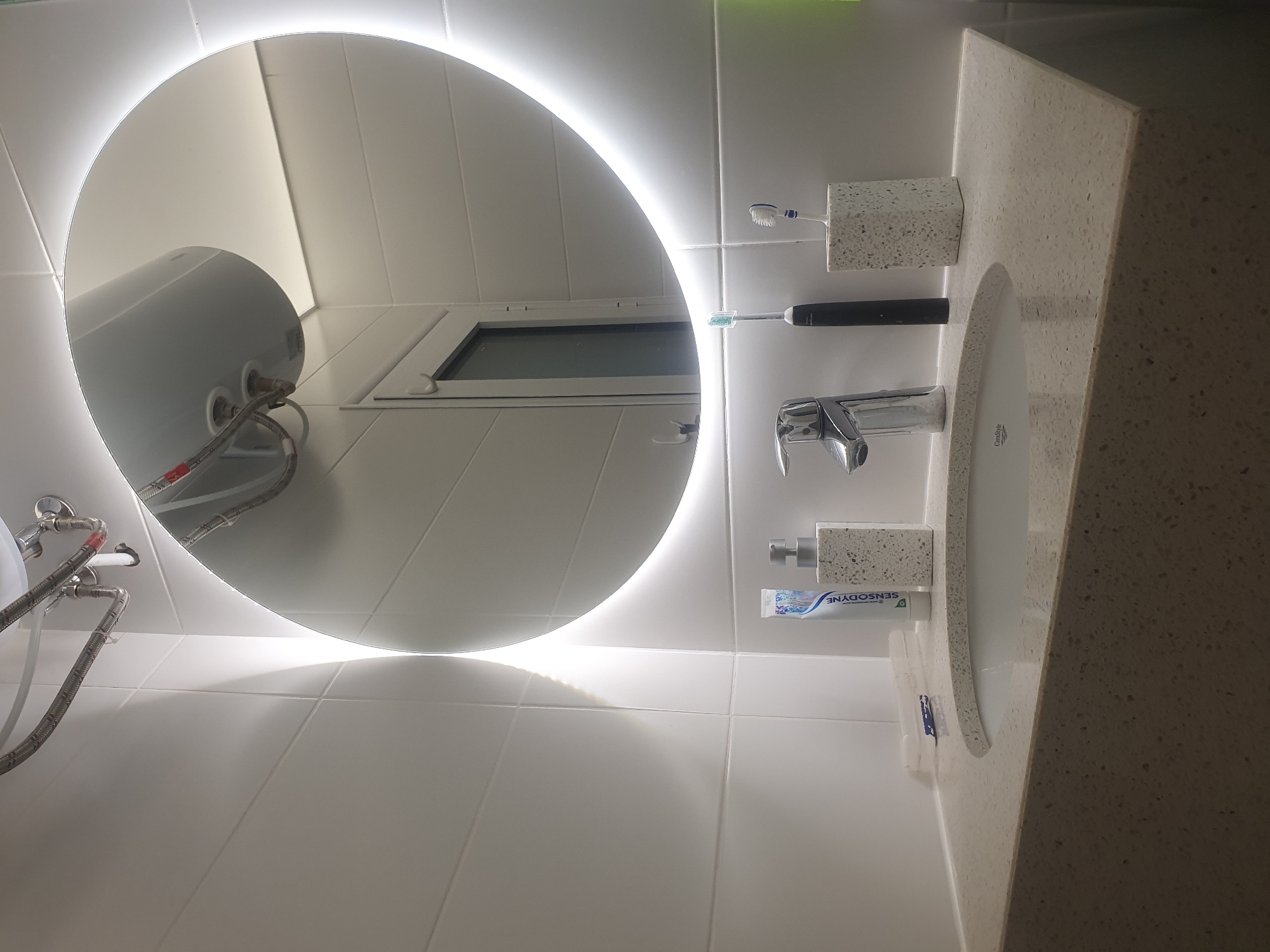 The image captures a bathroom interior, likely a residential space, featuring a circular, illuminated mirror reflecting elements of the room. Reflected in the mirror is a water heater and a window. The bathroom has white tiled walls. Next to the sink is a toothbrush holder with toothbrushes, a faucet, a soap dispenser, and a tube of Sensodyne toothpaste. The vanity top has a speckled beige pattern. The overall impression is of a clean, modern bathroom setting. The location, Plovdiv, Bulgaria, is not directly verifiable from the visual information in the image.