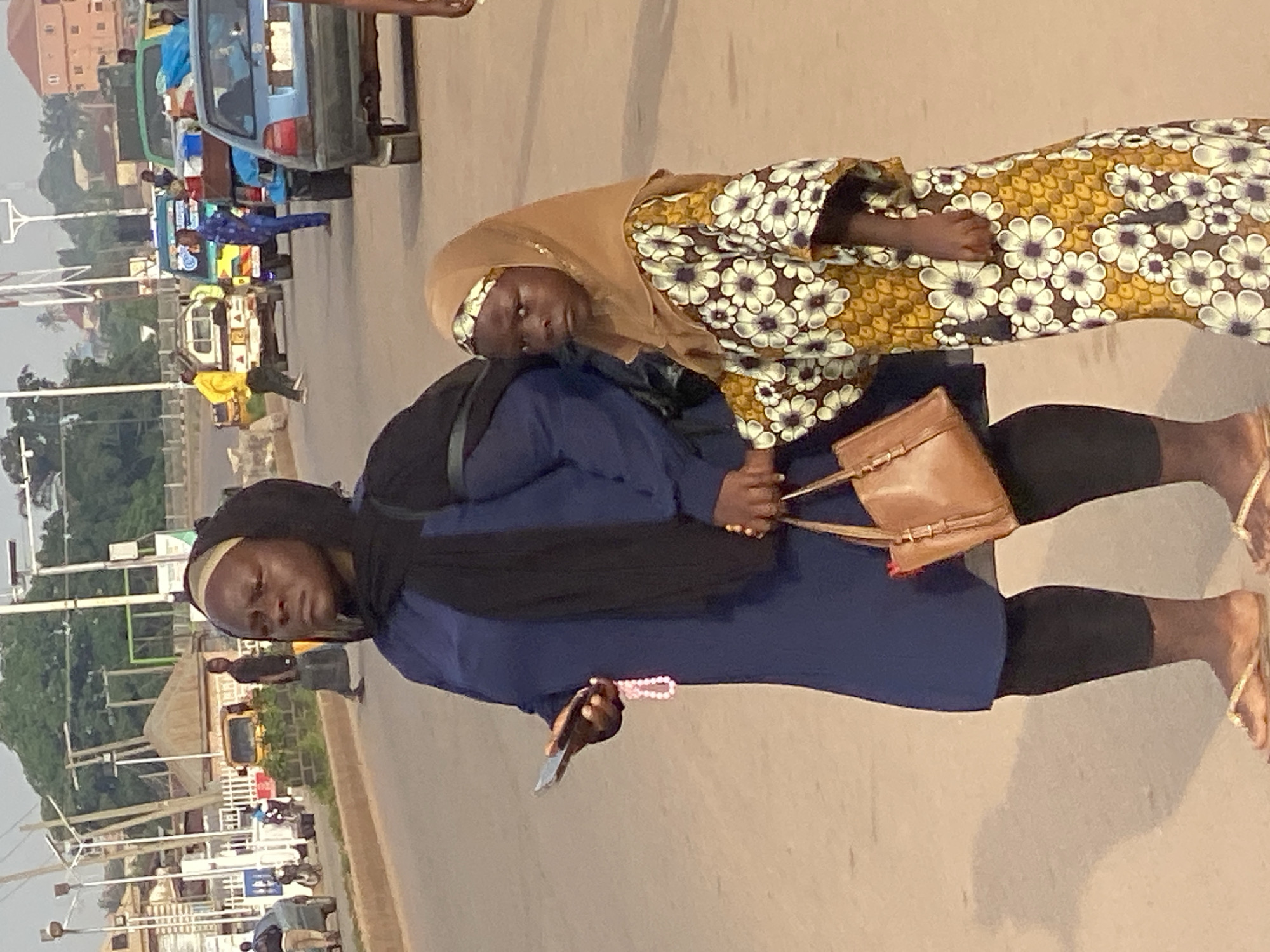 In Sabon Tasha, Nigeria, a woman in a blue dress and black headscarf holds a smartphone in her left hand, with a pink beaded bracelet dangling from her wrist. She also carries a brown handbag. Beside her stands a young girl wearing a light brown hijab and a long, patterned dress featuring white flowers with dark centers on a mustard-yellow background. The girl is holding the woman's hand. They are standing on a paved street, with other people and vehicles visible in the background. Cars, including a blue sedan and a green pickup truck loaded with luggage, are on the road, suggesting a busy transportation area. Buildings and utility poles are also present in the scene.