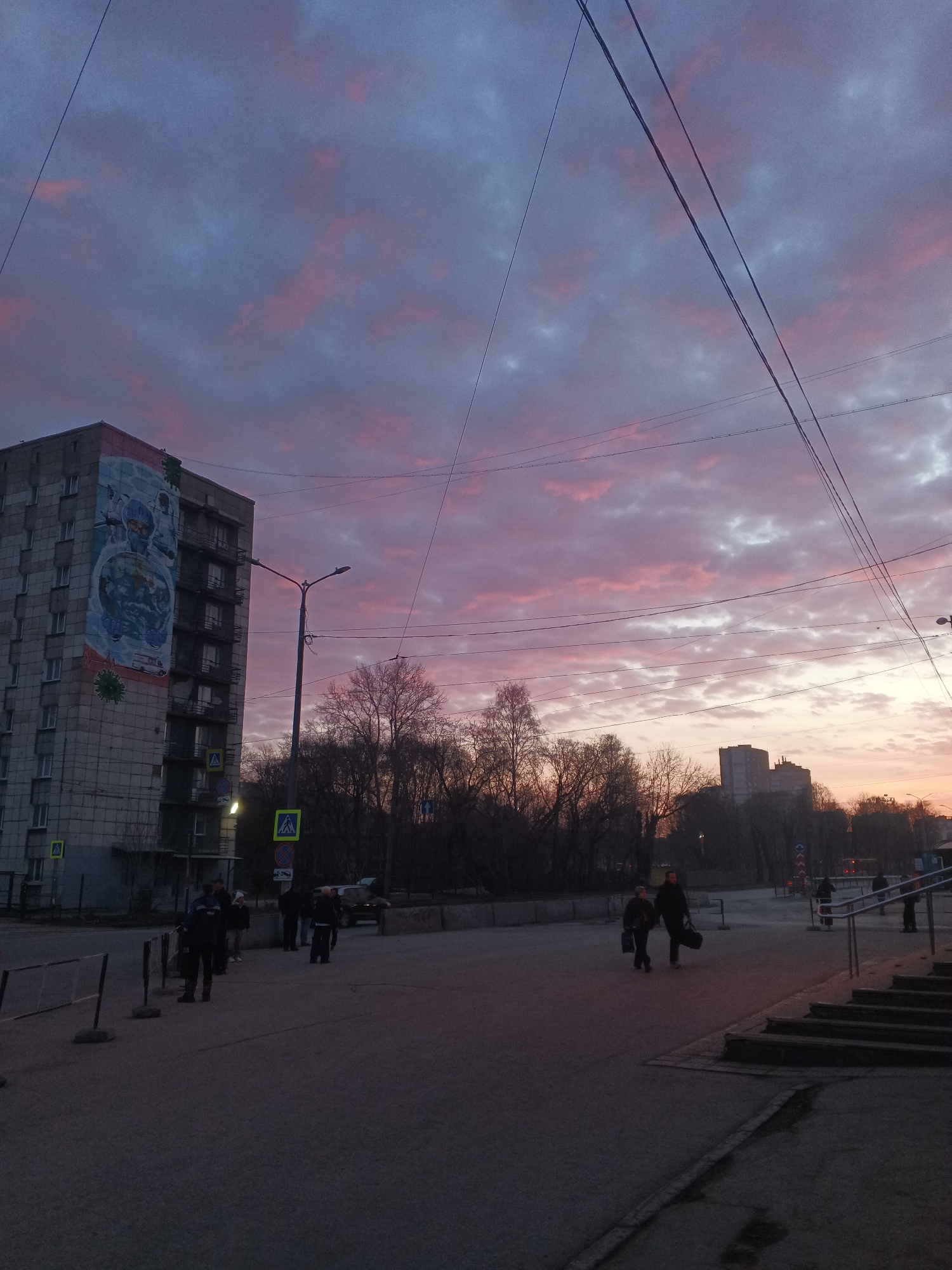 The image depicts a city street scene in Perm, Russia at dusk. A large, Soviet-era apartment building with a mural (appearing to be related to the COVID-19 pandemic) on its side stands to the left. Overhead power lines cross the sky, which is filled with pink and grey clouds. Several people are walking on the pavement, some in groups. There are trees and other buildings in the background. Streetlights are present, and a "zebra crossing" pedestrian sign is visible. Steps leading upwards are on the right side of the image.