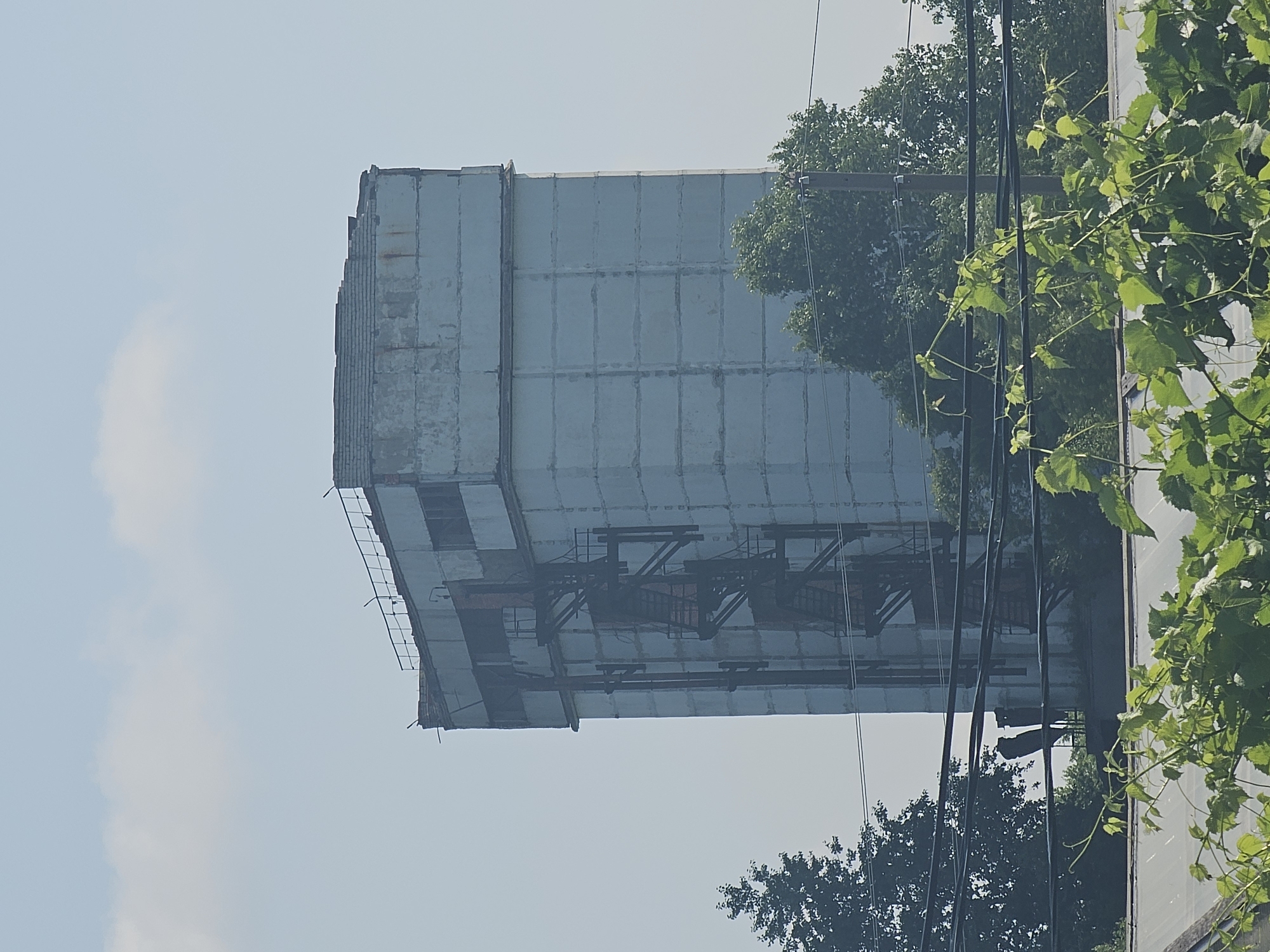 An aging, multi-story concrete structure with an exterior metal staircase stands against a hazy blue sky. The building appears weathered, with visible seams between the concrete panels and some signs of rust on the staircase. Trees and power lines are in the foreground and surrounding the base of the building, obscuring the ground level. The top of the building has a partially collapsed or damaged roofline. The overall scene suggests an industrial or abandoned site in Georgievka, Russia.