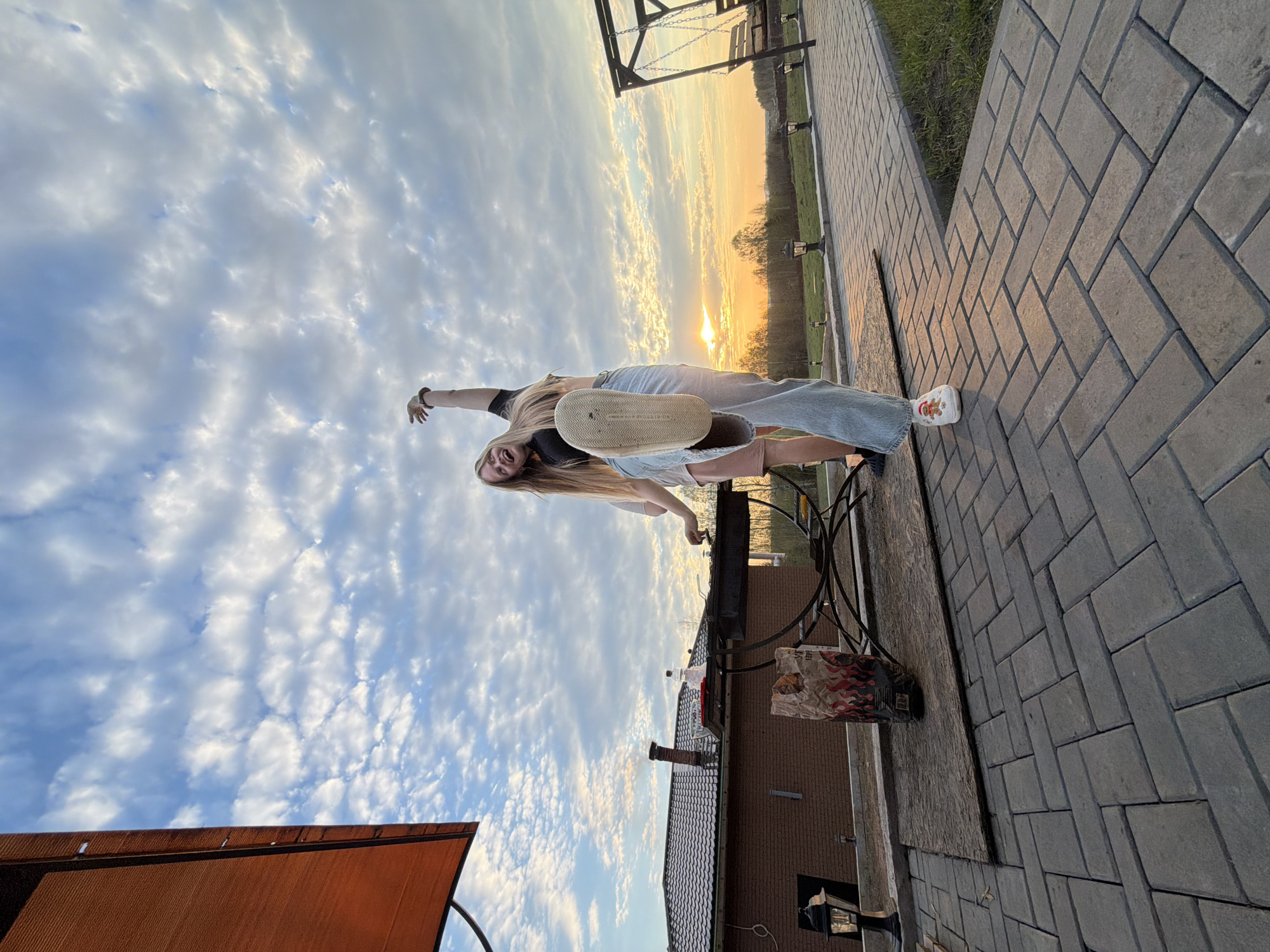 A young woman with long blonde hair, wearing a black top, blue jeans, and white sneakers with a colorful design, is lying on her back on a paved brick surface. Her left arm is extended towards the sky, while her right arm is bent, holding up a shoe sole. The sky is visible with numerous scattered clouds and the sun setting in the background. A brick building with a dark tile roof is visible in the lower left. A black metal table and grill are present to the right of the woman. Paved brick continues to the right, extending towards a distant swing set and green field. The location is likely outdoors in a residential area. Kamennoye, Russia, is not discernible from the image.