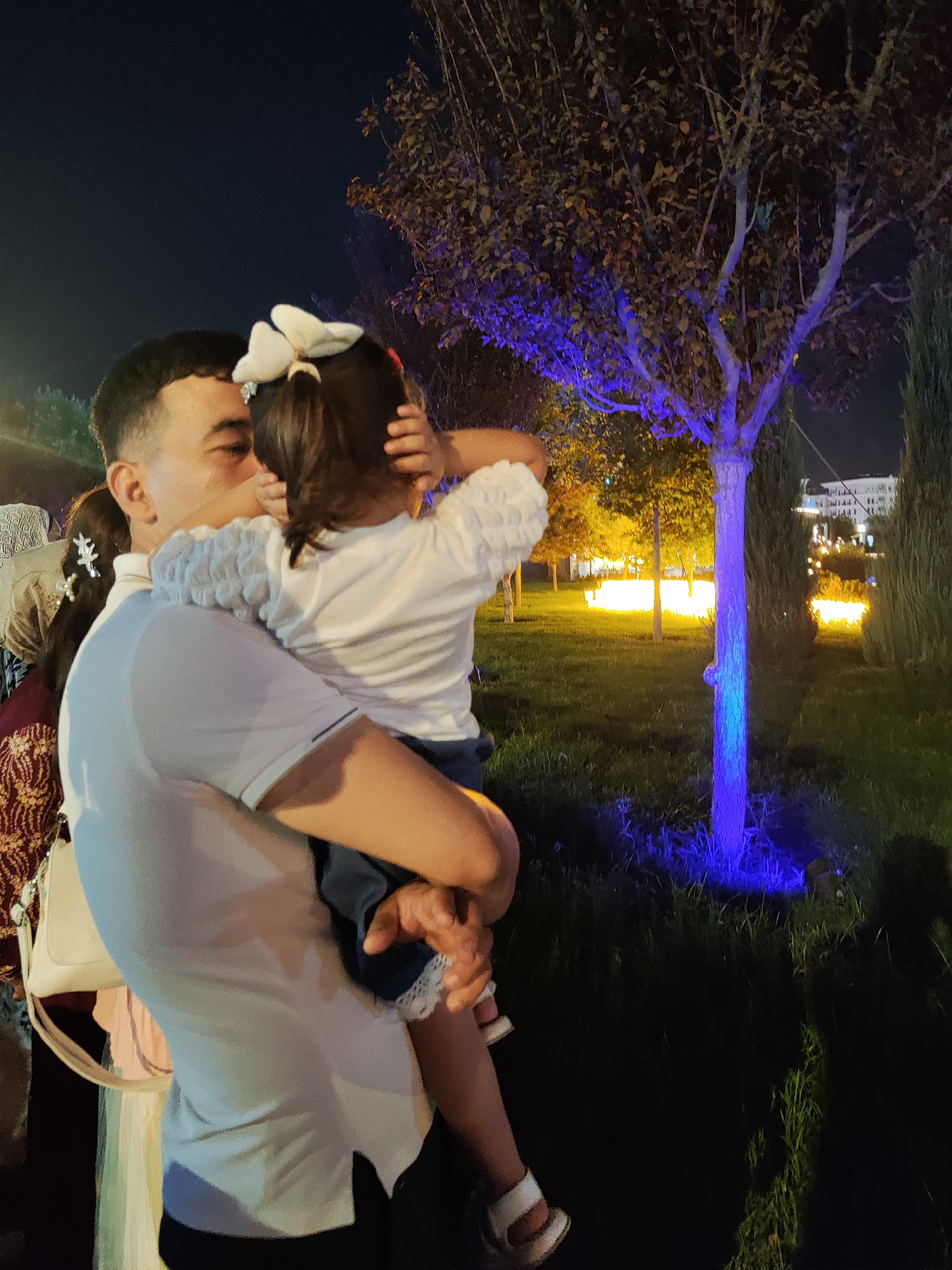 In this nighttime photograph, a man is holding a young child in his arms. The man is wearing a light blue polo shirt, and the child is wearing a white long-sleeved shirt and dark blue shorts. The child has a white bow in her hair. They appear to be in a park-like setting with trees and grass. One tree trunk in the background is illuminated with blue light. In the distance, there are lights from buildings and other trees, possibly indicating an urban area. The overall lighting suggests it is evening or night. The scene is likely a public space within the city of Tashkent, Uzbekistan, given the context provided in the prompt.