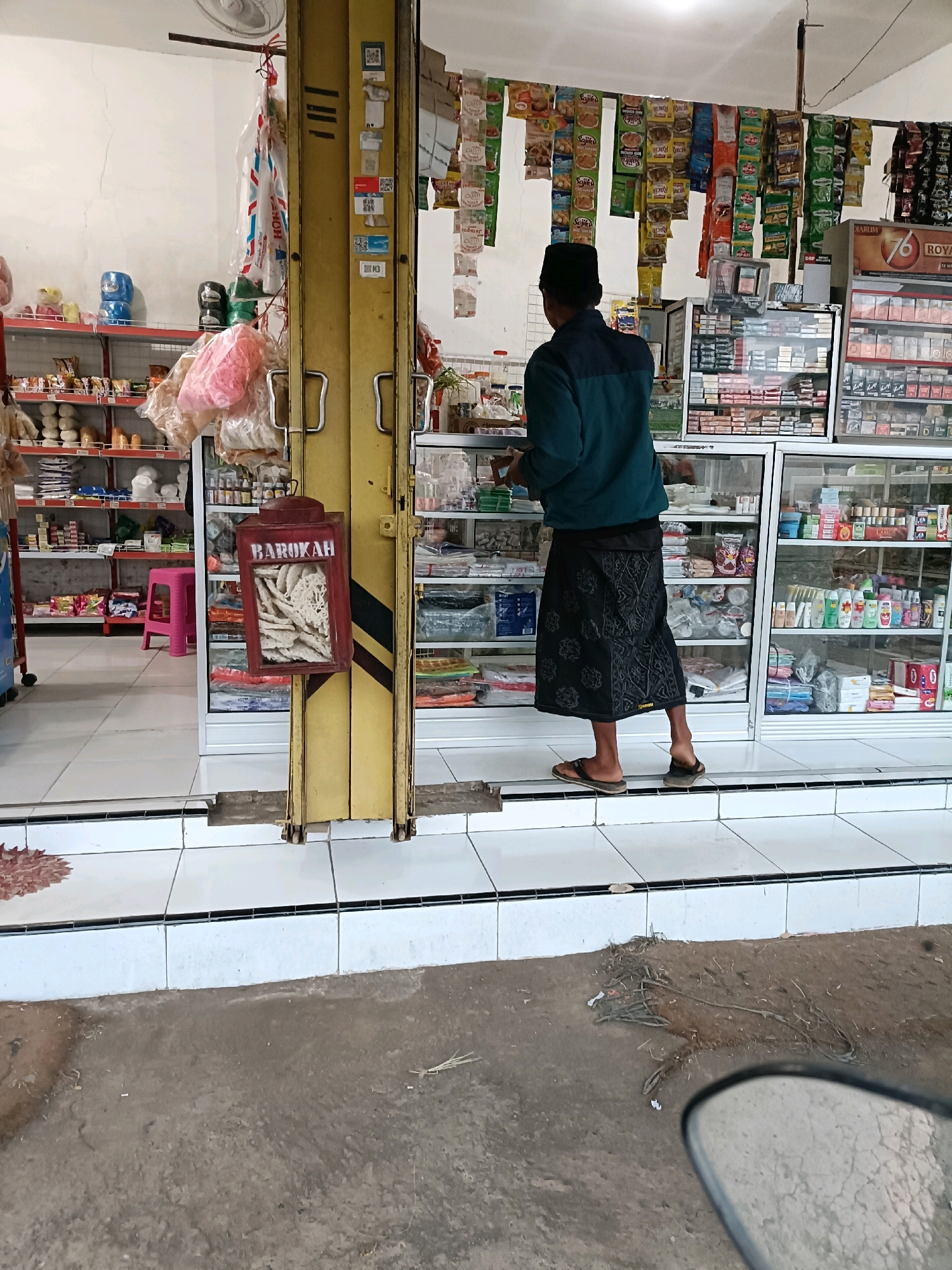 The image captures a street-level view of a small convenience store in Tamanan, Indonesia. A man wearing a dark blue long-sleeved shirt and a black sarong is standing in front of the store, facing away from the camera and towards the interior. He is wearing sandals. The store features numerous shelves stocked with various products, including packaged snacks, drinks, and toiletries. A prominent red box with "BAROKAH" written on it is hanging from a yellow pole to the left of the man. The store appears to have a yellow metal gate that is partially open. In the foreground, the ground is made of concrete tiles and dirt. The environment suggests a typical street scene in a residential or commercial area.