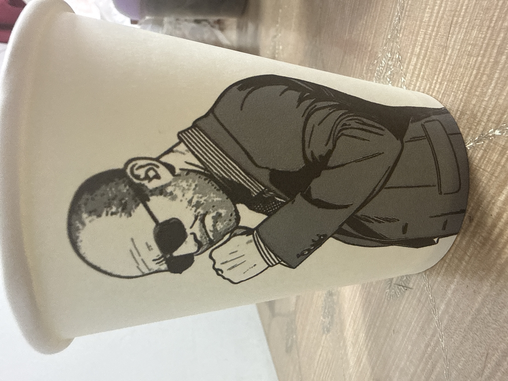 A close-up shot shows a disposable coffee cup with a black and white illustration of a bald man wearing sunglasses and a suit. The man is depicted from the chest up, with his right fist resting near his chin and his head tilted slightly down. The illustration has a textured, stippled appearance. The cup is light-colored and appears to be made of paper. It is placed on a wooden surface with a visible grain pattern. The background is out of focus, but suggests an indoor setting. There is no discernible information connecting the image or its contents to the city of Beslan, Russia.