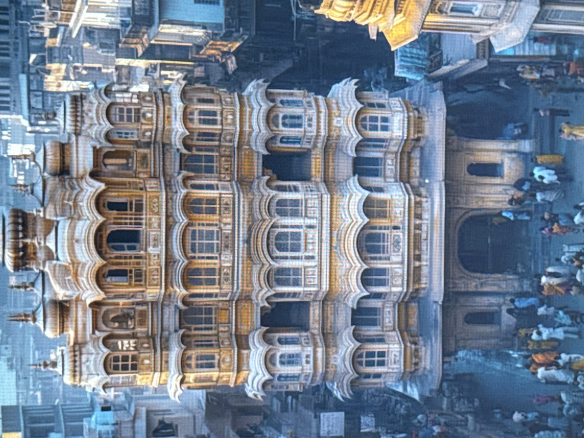 This image depicts a bustling street scene in front of an ornate, multi-story building with intricate architectural details, including numerous arched windows and balconies. The building appears to be made of a light-colored stone with some golden accents, suggesting a rich historical or cultural significance. The perspective of the image is from an elevated viewpoint, looking down at the scene.
In the foreground, the ornate building dominates the frame. The repetition of arched windows and decorative elements creates a sense of grandeur and complexity. Behind the building, a crowd of people is visible, dressed in various colorful clothing, suggesting a lively atmosphere. They appear to be walking or standing, engaged in daily activities. The presence of many people and the colorful attire hint at a public gathering or a busy market area.
The background shows more buildings and structures, indicating an urban setting. The overall impression is one of a vibrant and densely populated location. The lighting suggests it could be daytime, possibly late afternoon given the warm tones on the building. There are no clear indications of the specific time of day or weather conditions, though the scene appears well-lit. No visible text is present in the image. The scene is rich with architectural detail and human activity.