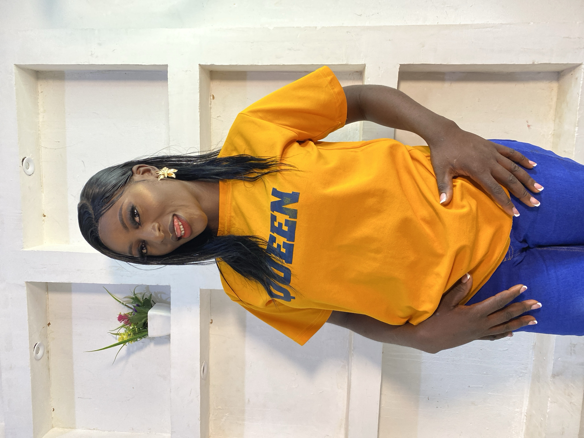 A medium shot shows a Black woman posing in front of a white, recessed wall with decorative niches. The woman has long, dark hair and is wearing a bright orange t-shirt with the word "QUEEN" printed in blue letters across the chest. She is also wearing blue jeans. She has a gold earring shaped like a flower in her right ear and is sticking her tongue out playfully. Her hands are clasped in front of her. In one of the niches behind her, there is a small white planter with artificial flowers. The overall scene is well-lit.