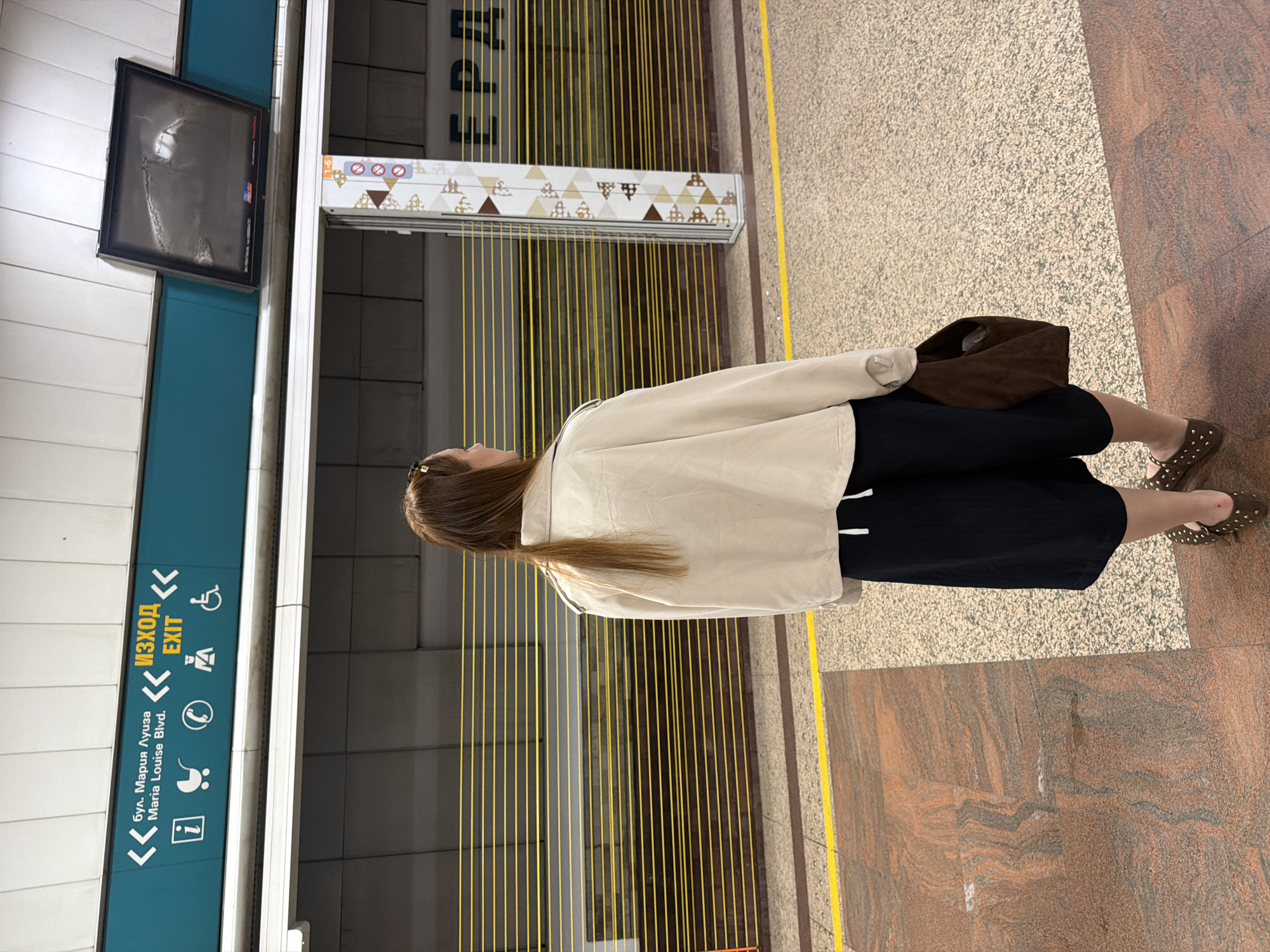 A woman is standing in what appears to be the Sofia Metro. She is wearing a beige jacket, black pants, and brown shoes with studs. She also has a brown handbag. The Metro station has signage indicating "Exit" in both Bulgarian and English, and lists "Maria Louise Blvd." in Bulgarian and English. The station has a digital display. The walls have decorative vertical yellow lines. The floor is patterned.