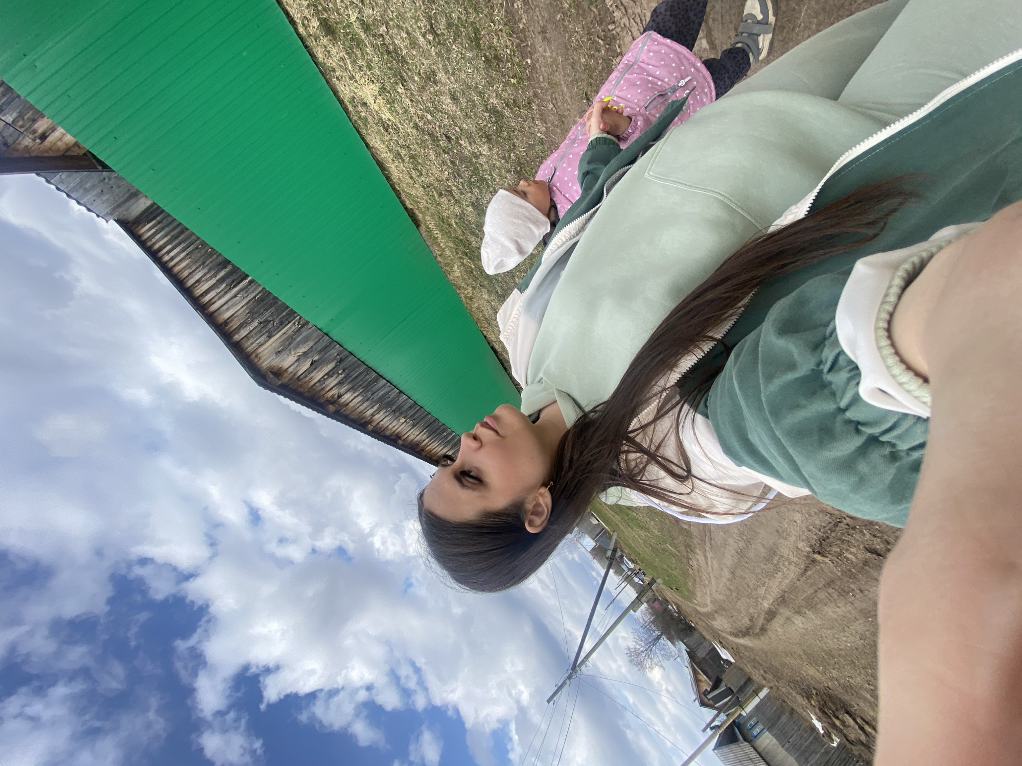 The image depicts a woman taking a selfie outdoors in Zhigalki, Russia. She is wearing a light green sweatshirt and has long, dark hair. A child in a pink jacket and white head covering is lying on the ground next to her. A green-roofed building is visible in the background, along with a blue sky with clouds. A dirt road and other buildings are also visible in the distance.