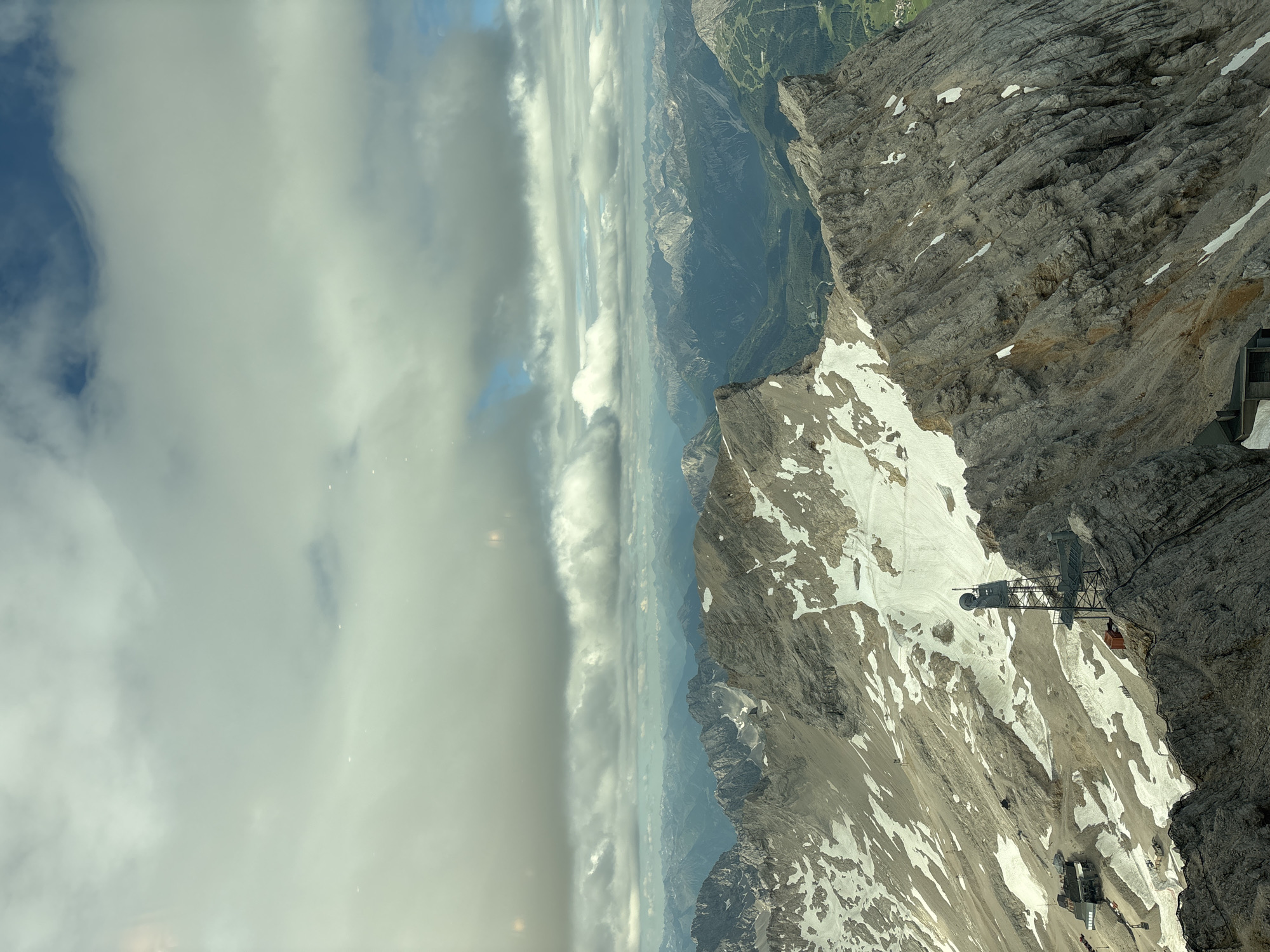The image depicts a panoramic view of a mountainous region, likely taken from a high vantage point. Jagged, rocky peaks dominate the foreground and midground, with patches of snow scattered across their slopes. In the distance, a vast expanse of layered mountains fades into a hazy blue horizon. The sky is filled with a dramatic arrangement of clouds, ranging from dense, grey masses to thinner, wispy formations that allow glimpses of blue sky.
Specific structures are visible on the mountainside. To the left, a complex of buildings, possibly a resort or research facility, is nestled into the terrain. A prominent metal tower with a dish antenna stands on a rocky outcrop in the mid-right of the frame, suggesting a communication or observation post. The ruggedness of the mountains suggests a high-altitude, alpine environment. While the image is attributed to Ehrwald, Austria, specific landmarks within Ehrwald itself are not definitively identifiable from this perspective. The scene is devoid of any visible human figures or active events.
