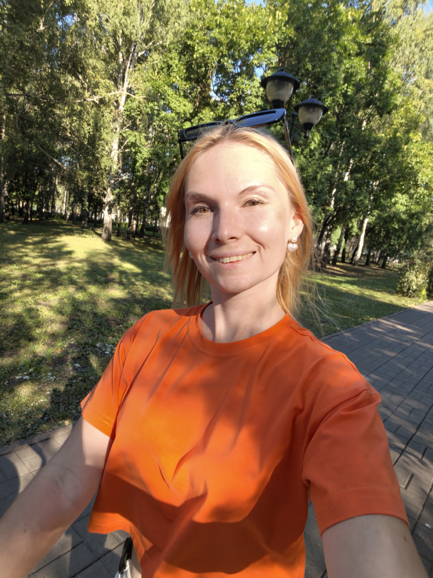 A young woman with blonde hair and fair skin is taking a selfie outdoors in a park. She is wearing an orange t-shirt and a pair of heart-shaped pearl earrings. Her sunglasses are perched on top of her head. The background consists of green trees and a paved path, with sunlight dappling through the leaves. The overall scene suggests a pleasant, sunny day in an urban park, possibly in Sterlitamak, Russia.