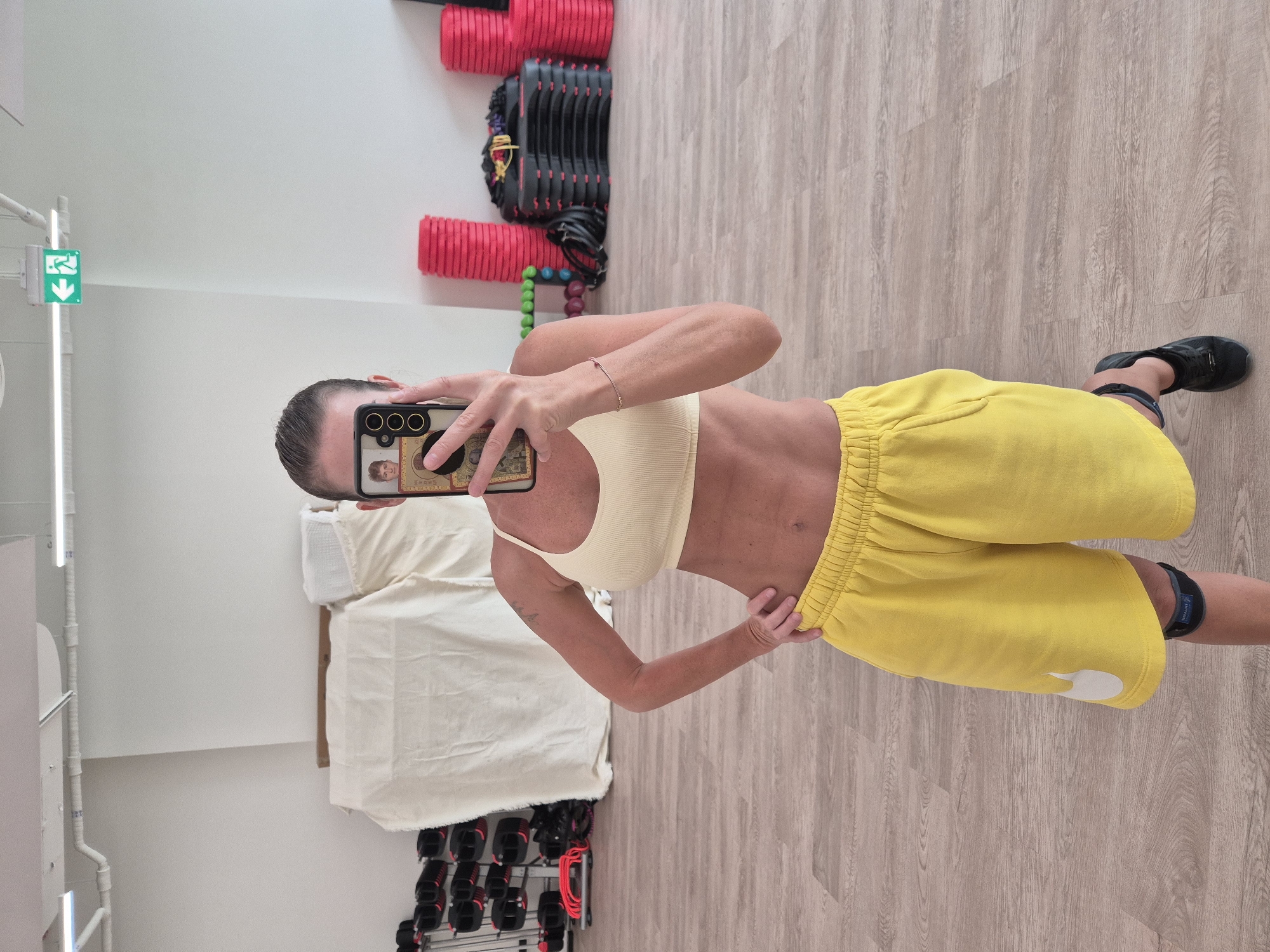 A woman is captured in a mirror selfie, standing in what appears to be a gym or fitness studio. She is wearing a light yellow ribbed sports bra or crop top and bright yellow athletic shorts with an elastic waistband. Her hair is pulled back, and she holds a smartphone with a decorative case in her right hand, covering her face. Her left hand rests on her hip, and a thin bracelet adorns her left wrist. On her left calf, a black strap or device with the word "TREXIAM" faintly visible is present. She is wearing black athletic shoes.
The setting features light-colored wooden or laminate flooring. In the background to her left, there's a white wall with a green emergency exit sign, depicting a running person and an arrow pointing down-left. Vertical light fixtures or pipes are also visible on this wall. Below, a light-colored fabric or towel is draped over what might be exercise equipment or a bench. To her right, a rack holds various dumbbells or weights, some red and some black, stacked on shelves. Above these, red ribbed fitness tubes or rollers are arranged, and below, a row of colorful beaded objects can be seen. The overall lighting suggests an indoor, well-lit environment, likely during the day.