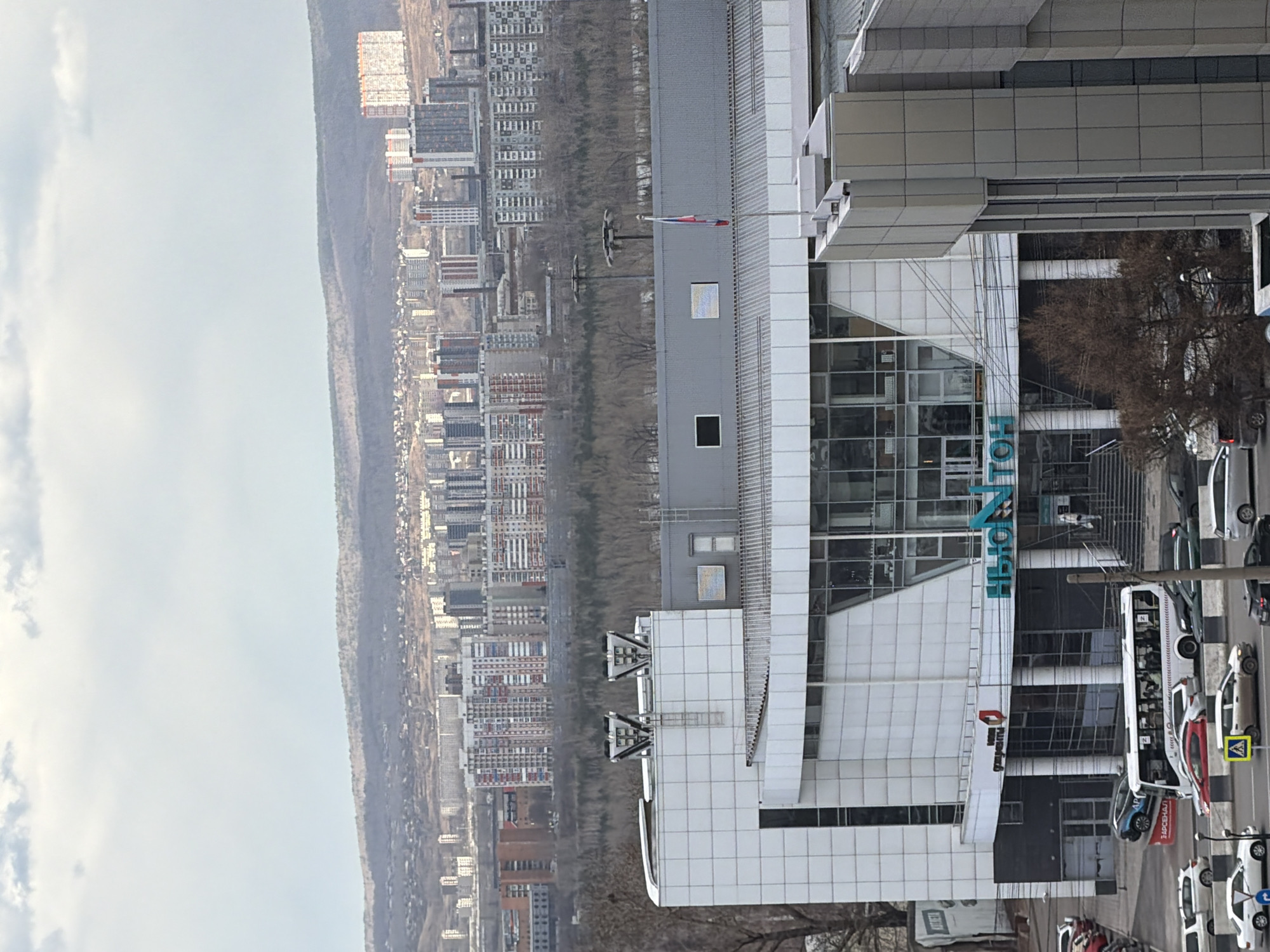 The image displays an urban landscape in Krasnoyarsk, Russia, seen from an elevated perspective on an overcast day. In the foreground, a modern multi-story building dominates the right side of the frame. This structure features a contemporary design with white and grey paneled facades and large glass windows. On its roof, several technical installations are visible, along with a flagpole flying a flag displaying red, white, and blue stripes, indicative of the Russian flag.
Below the foreground building, a street is visible with several parked cars and a white public transit bus. A yellow pedestrian crossing sign stands near the street. Further back, bare trees suggest the season is late autumn, winter, or early spring. The midground and background are filled with numerous residential apartment buildings, forming a sprawling cityscape that extends to a distant, wooded ridge.
No specific dynamic activity is taking place; the scene captures a static moment of city life. The time of day is daylight, under a cloudy sky.
Visible text on the main foreground building includes large blue letters spelling "ЭЛЕКТРОН" (ELECTRON). Below this, a red logo with white text, likely "Сбербанка" (Sberbank), can be seen. Another red sign near the street reads "ТАРСЕНАЛ" (TARSENAL).
