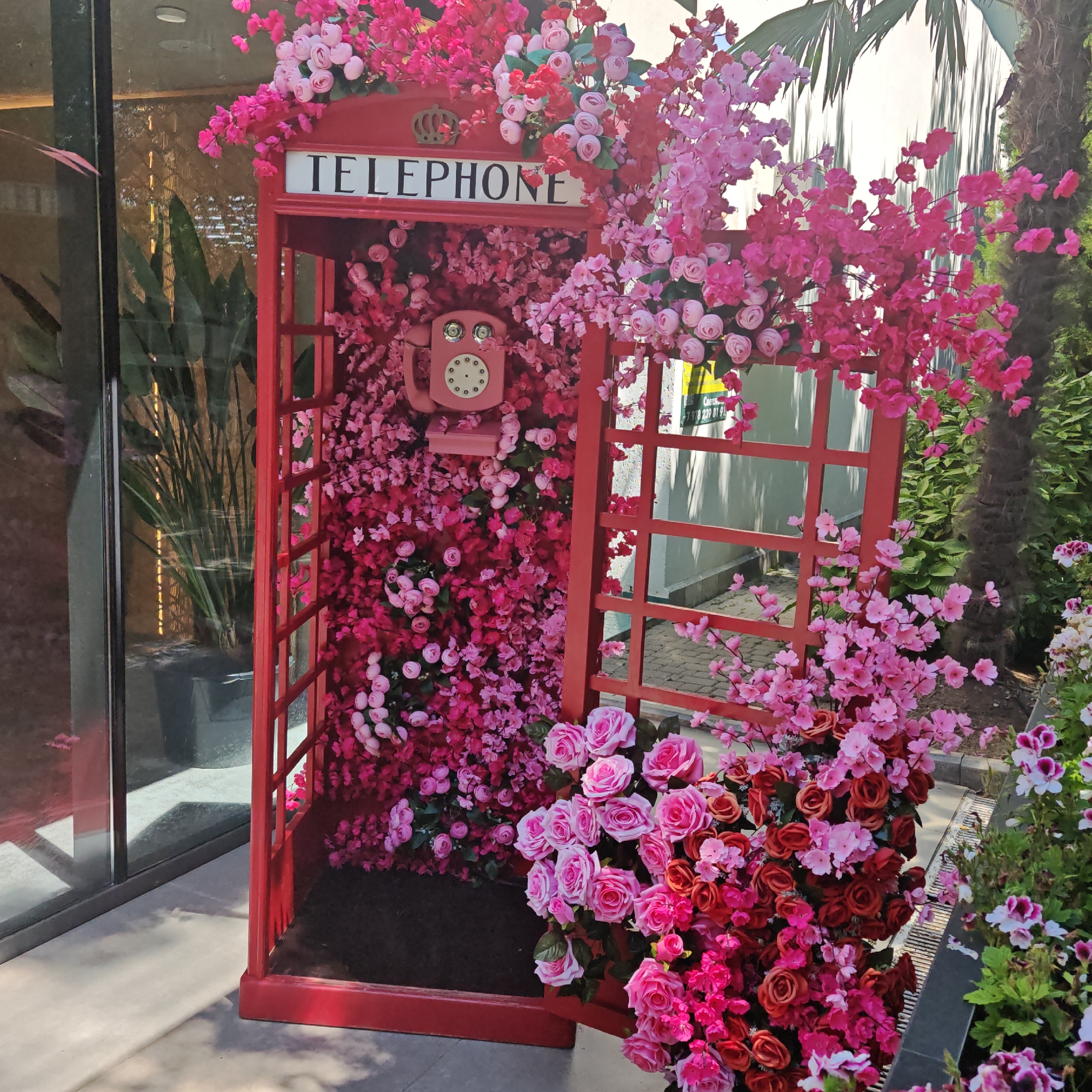 An outdoor scene features a red, vintage-style British telephone booth overflowing with pink and red artificial flowers, creating a vibrant and decorative display. The telephone booth is adorned with the word "TELEPHONE" in white lettering on a black background. Inside the booth, a pink vintage rotary phone is visible, also surrounded by flowers. The booth is placed on a grey paved surface, with greenery and a palm tree visible in the background. Sunlight casts shadows on the ground, indicating an outdoor setting during the daytime.