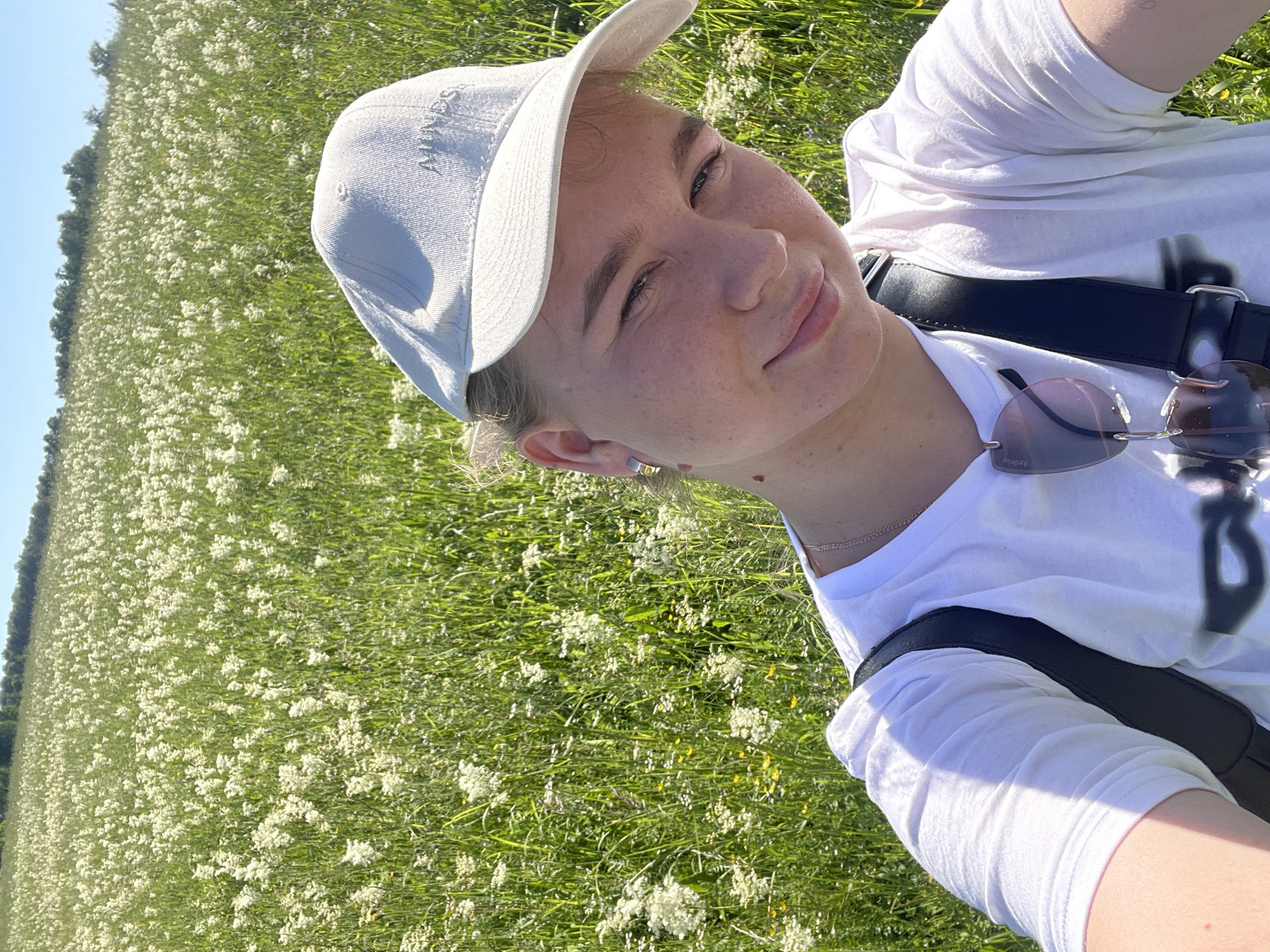 The image is a selfie taken in a grassy field with white flowers. A young person, wearing a white t-shirt, a white baseball cap, and sunglasses hanging from their shirt, is smiling at the camera. They have blonde hair and are wearing a silver necklace. The background is a blurry expanse of green grass and white flowers, suggesting an outdoor, natural setting. The image appears to be taken on a sunny day.