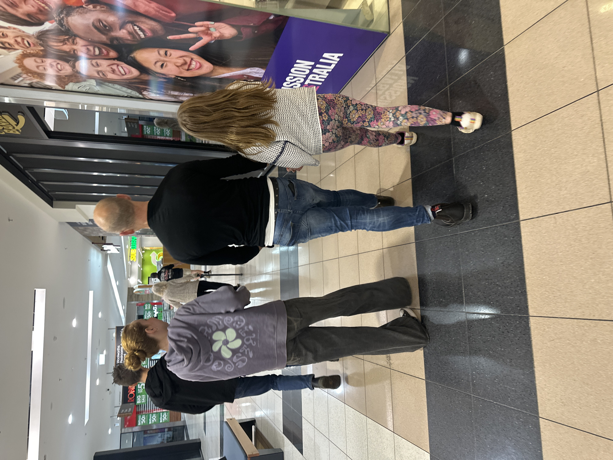 A group of four people are walking through a shopping center. The group consists of two adults and two children. The two adults appear to be a man and a woman, and the two children are also a boy and a girl. They are all walking away from the camera, and their faces are not visible. The shopping center appears to be indoors, with a tiled floor and fluorescent lighting. There are shops visible in the background, including a Subway and a Robin Hood. A large poster of smiling faces is also visible on the right side of the image. The people are dressed in casual clothing. The man in the foreground is wearing a black t-shirt and blue jeans. The woman is wearing a purple hoodie and grey pants. The boy is wearing a black hoodie and blue jeans. The girl is wearing a white and black patterned sweater and floral leggings. They are walking on a section of the floor that is tiled with a darker shade of tile, bordered by a lighter shade of tile. The context provided by the poster and the store names suggests this location is likely toormina, Australia.
