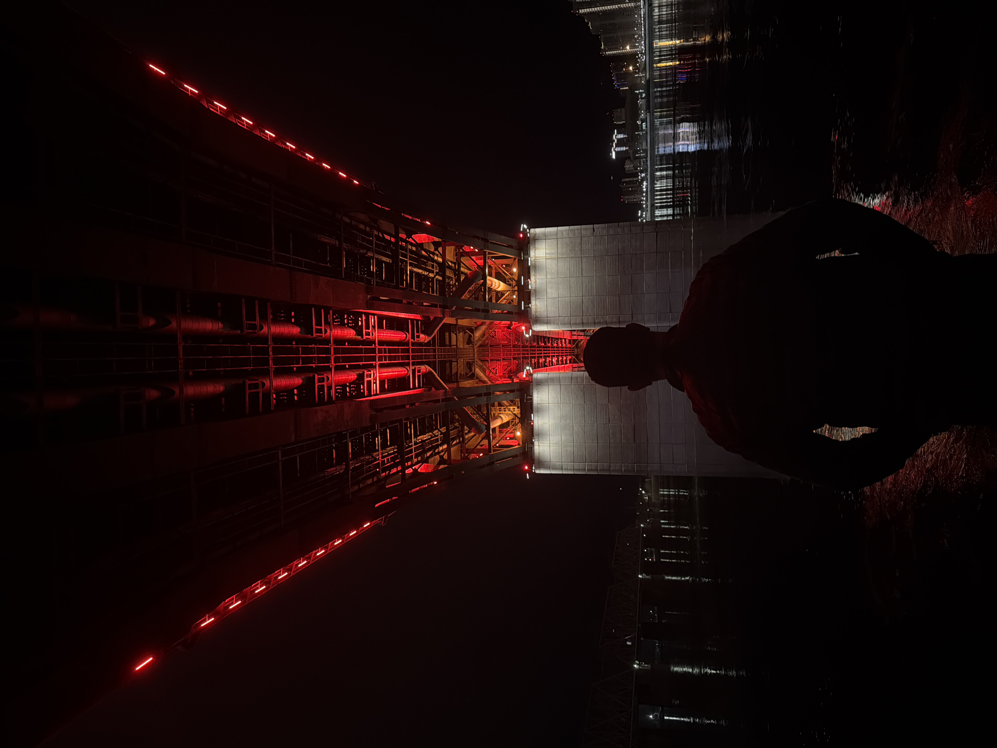 The image depicts a night scene in Krasnoyarsk, Russia, featuring the Krasnoyarsk Metallurgical Plant with its red lighting reflected in a body of water. The reflection creates a symmetrical composition, with the plant structure visible on the left side of the image. A dark silhouette of a person is visible on the right side, partially obscuring the building reflected in the water. In the background, the lights of nearby buildings and structures can be seen, further reflecting in the water. The overall tone is dark and industrial, with the red lighting of the plant creating a striking contrast against the night sky.