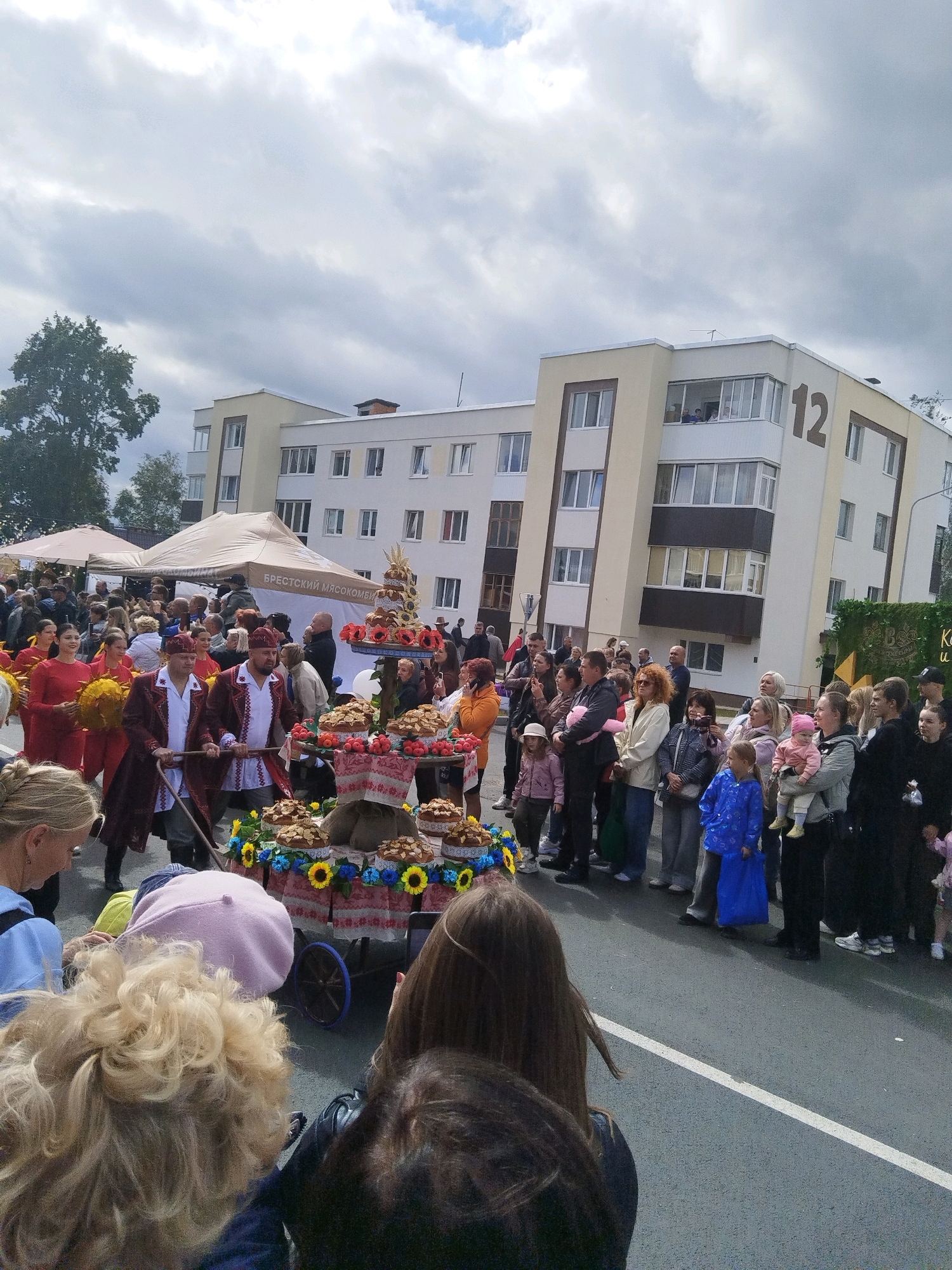 In an outdoor urban setting, a crowd of people gathers to observe a procession. A building with a large "12" visible on its facade stands in the background. In the foreground, a cart decorated with floral arrangements and baked goods is being pulled by two individuals in traditional attire. Spectators of various ages line the street, some holding children. The sky is overcast.