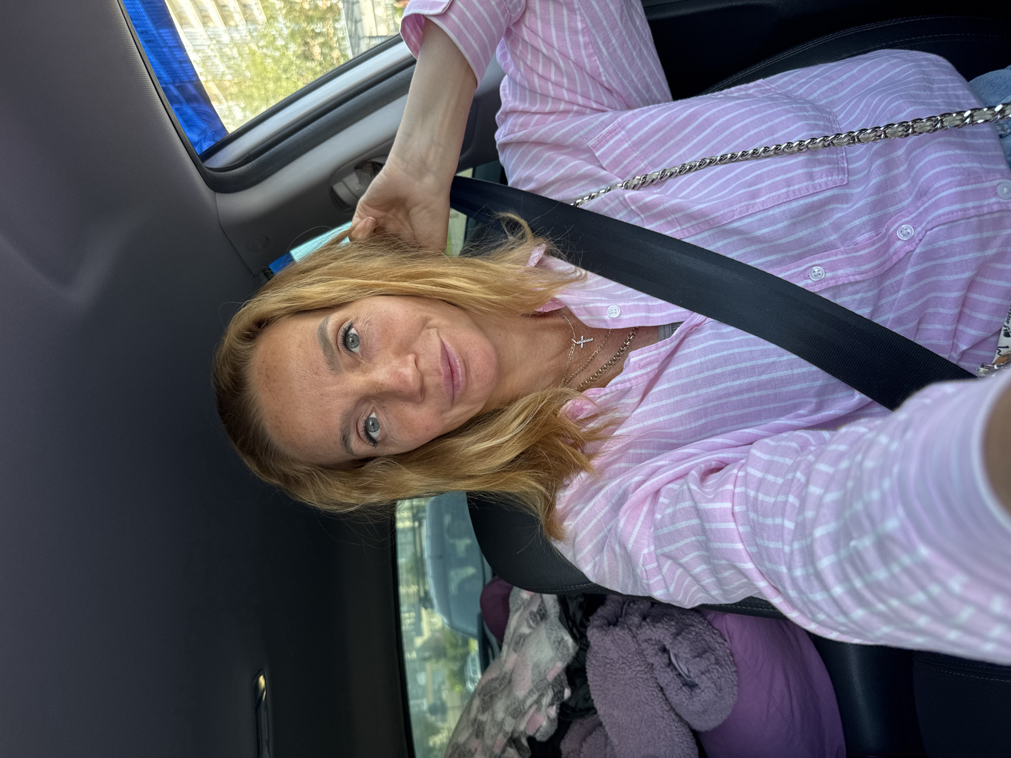 A woman with blonde hair is sitting in the driver's seat of a car, wearing a pink and white striped button-down shirt and a silver necklace with a cross pendant. She is looking at the camera and touching her hair. A black seatbelt is fastened across her chest. In the background, there are folded blankets and pillows in the passenger seat, and through the windows, a city street with buildings and trees is visible. This scene is likely in Saint Petersburg, Russia, given the architectural style of the buildings and the presence of a car, common elements in urban Russian environments.