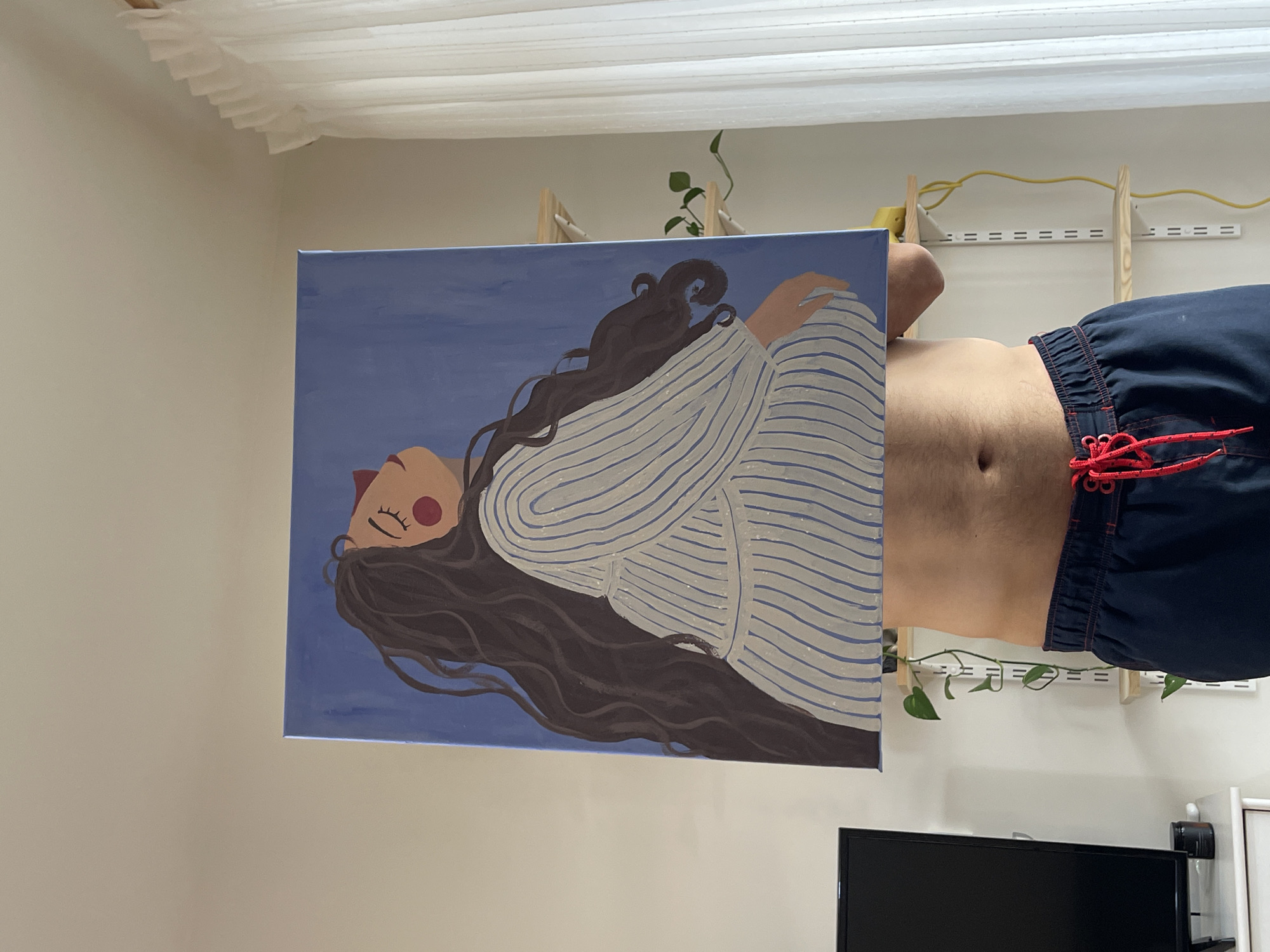 The image shows a person holding a painting in front of their torso. The painting is of a woman lying down, with long brown hair and wearing a striped shirt. The person is wearing blue shorts with a red drawstring. The background includes a white wall, a white curtain, a shelf with plants, and a television. The location is unknown.
