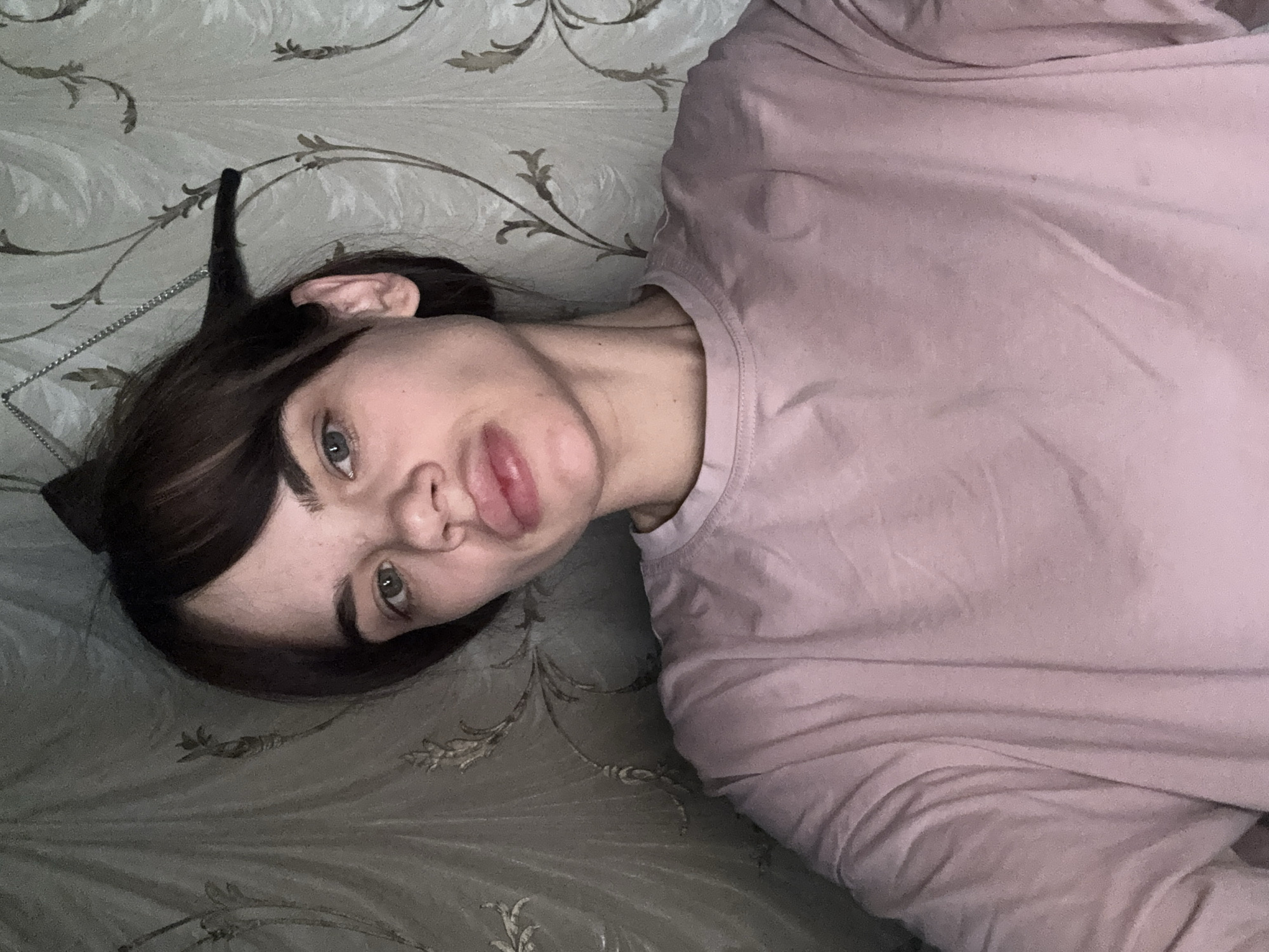 A person with dark hair and fair skin is lying on a patterned surface. The person is wearing a pink long-sleeved shirt and appears to be looking at the camera with a slightly pouting expression. A black object resembling a cat ear is visible near the person's hair. The background has a floral pattern. There is no information available to determine if the image was taken in Rostov-on-Don, Russia, or to indicate the location of the subject.