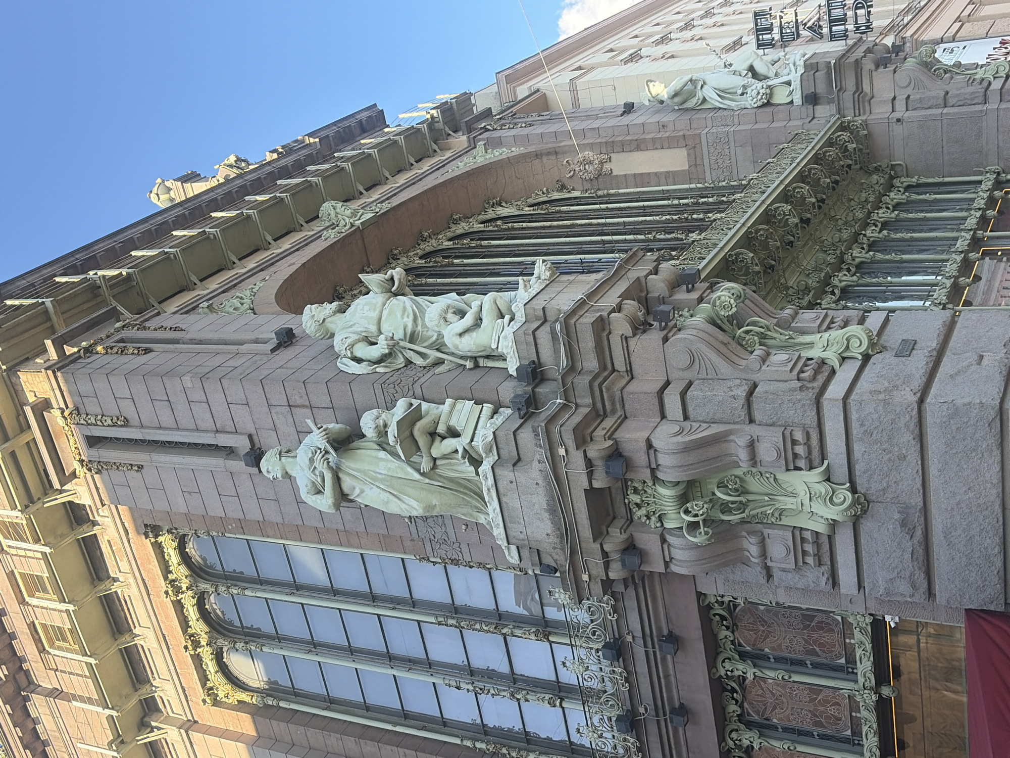 The image shows the Singer House, a historic Art Nouveau building located on Nevsky Prospekt in Saint Petersburg, Russia. The building features ornate architectural details, including large arched windows, balconies with intricate ironwork, and decorative sculptures. Two prominent statues, a woman with a book and a child reading, are visible on the corner of the building. The sky is clear blue, indicating a sunny day. A red awning is partially visible at the bottom left of the image.