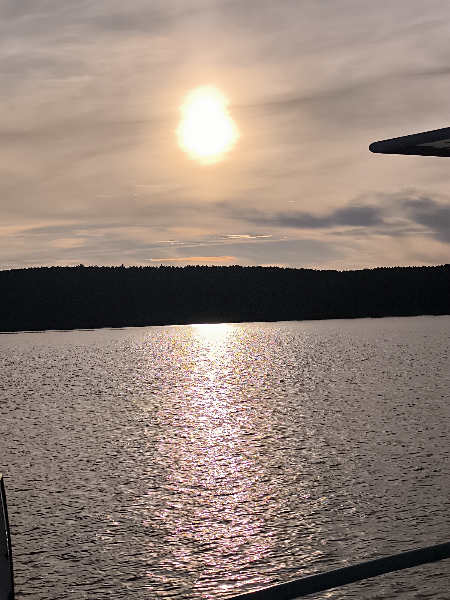 An expansive, calm body of water reflects the muted light of a sun partially obscured by clouds. The horizon is marked by a dense, dark silhouette of a forest against a pale, peachy sky. A bright, diffused sun is visible in the upper center of the frame, casting a shimmering, colorful path across the water's surface. The lower portion of the image shows the edge of what appears to be a boat or dock. The scene evokes a sense of peaceful twilight or dawn, likely captured from a vantage point on the water. This image is consistent with landscapes found along the Curonian Lagoon in Neringa, Lithuania, particularly during the softer light conditions of sunrise or sunset, where the water and forest edge are prominent features.