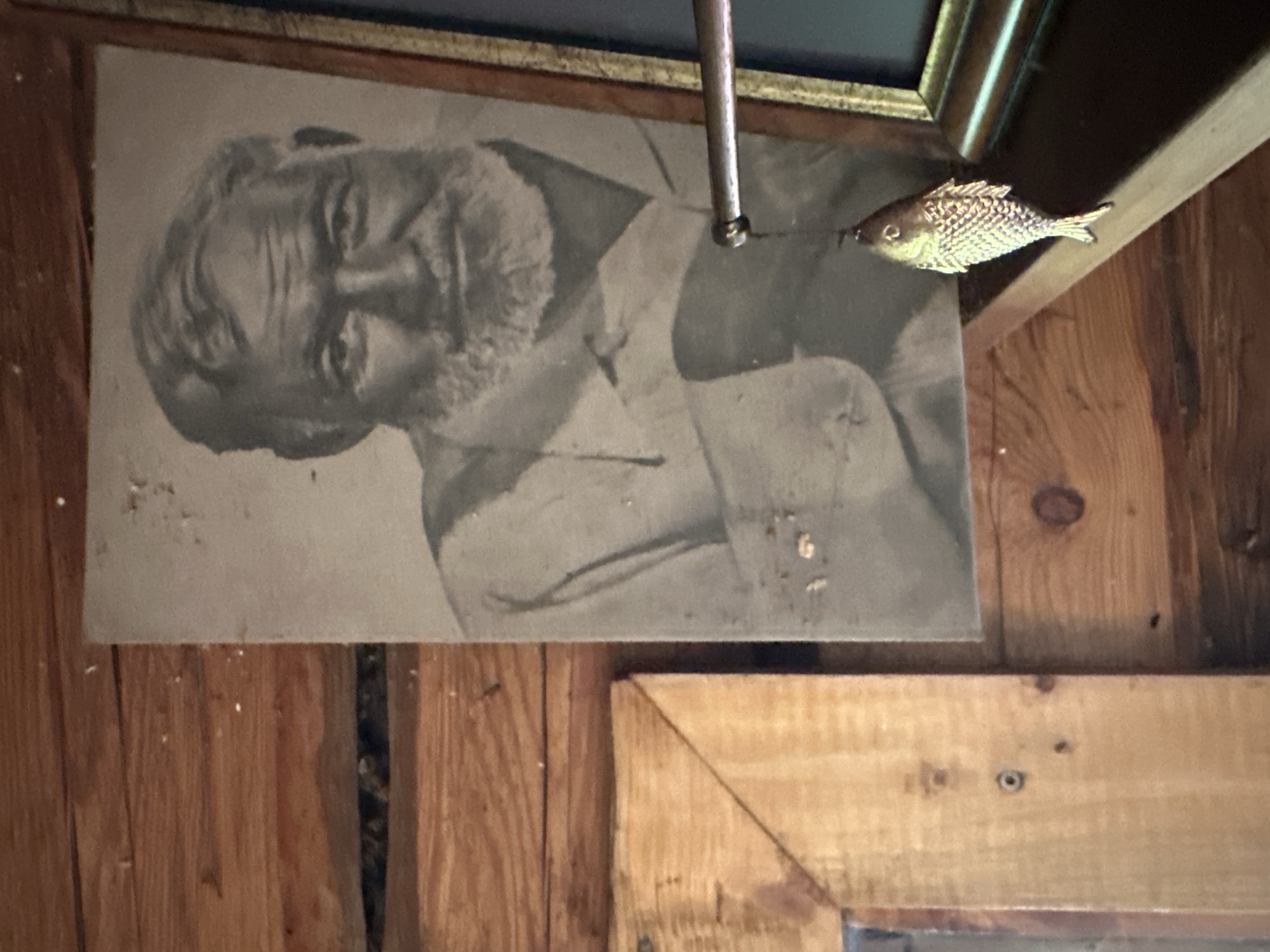 The photograph depicts a black and white portrait of an elderly man with a beard, identified as Ernest Hemingway. The portrait is affixed to a wooden wall with visible planks and knots, characteristic of a rustic interior. A decorative brass fish charm hangs from a thin chain attached to what appears to be a thin rod or wire, casting a shadow on the portrait. To the left of the portrait, a corner of a wooden frame is visible, also mounted on the wooden wall. The overall setting suggests a humble, perhaps historical, dwelling in Norviliškės, Lithuania, possibly within the Norviliškės Manor or a related structure. The lighting is subdued, emphasizing the textures of the wood and the aged quality of the photograph.