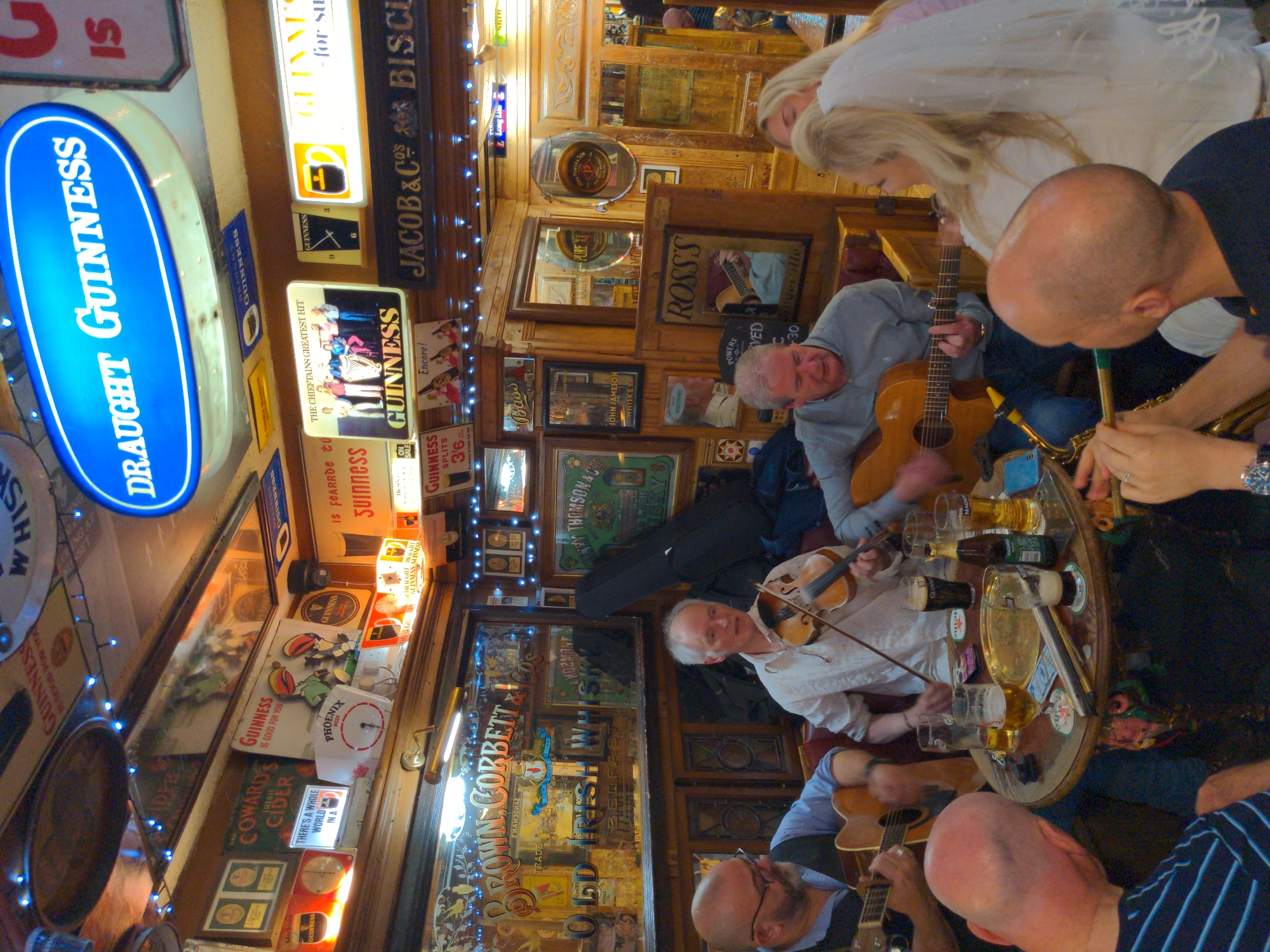 The image depicts an interior scene of what appears to be a traditional Irish pub. The walls are adorned with numerous vintage signs advertising brands such as Guinness, Old Irish Whiskey, and John Jameson. Wooden paneling and furniture contribute to the rustic ambiance. In the foreground, a group of people are gathered around a circular table. A man on the left is playing an acoustic guitar. To his right, another man is playing the violin. A woman with blonde hair, possibly wearing a veil, is seated on the right. There are drinks on the table, including glasses of beer and a bottle. The overall impression is of a lively gathering, possibly a musical performance, within a historic establishment. The mention of Belfast, United Kingdom, is not directly evident from the visual information within the image itself, but the presence of Irish-themed advertisements and the general aesthetic strongly suggest a location within Ireland, and Belfast is a plausible setting for such a pub.