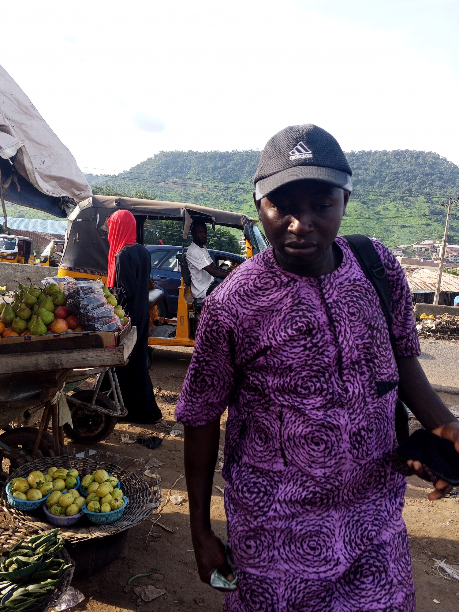 A medium shot captures a man in a purple patterned kaftan and a black Adidas baseball cap standing in an outdoor market. He is looking down and slightly to his right. He carries a black backpack over his left shoulder and holds a dark smartphone in his right hand. Behind him, a fruit stand displays pears and other fruits, with a woman in a red headscarf and black dress standing near it. A yellow auto-rickshaw, likely a 'Keke Napep', is parked to the right of the fruit stand, with a man driving it. In the background, a green, wooded hillside rises above a town with buildings. The scene is consistent with a street market in Lokoja, Nigeria, a city known for its hilly terrain and vibrant street commerce. The ground is dusty earth, typical of the area. The lighting suggests it is daytime, with bright sunlight creating shadows.