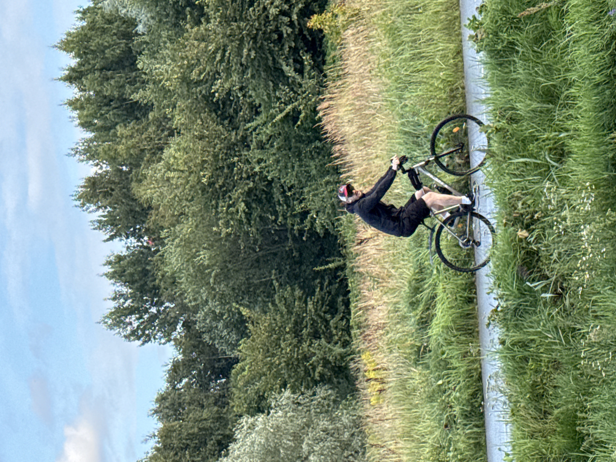 A person wearing a black hooded jacket and black shorts is cycling on a paved path. The person is also wearing a red and white baseball cap. They are riding a bicycle with black tires and a silver frame. The path runs alongside a field of tall green grass and wildflowers. In the background, there is a dense forest with various shades of green trees. The sky is blue with some scattered white clouds. The image appears to be taken in an outdoor, natural setting, possibly a park or a nature trail within or on the outskirts of Tallinn, Estonia.