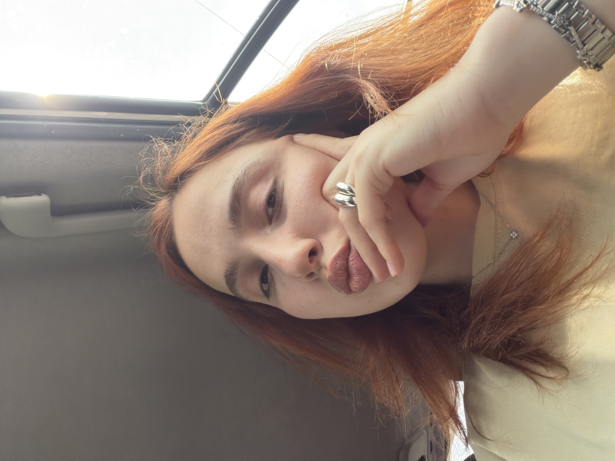 A selfie-style photograph depicts a young woman with auburn hair and fair skin. She is positioned within a car, visible through the sunroof at the top of the frame. The woman's face is partially obscured by her hand, which is adorned with a double-band silver ring. She wears a metallic wristwatch on her wrist and a delicate necklace with a small pendant. The backdrop includes the car's interior with grey upholstery. No information is available to determine the location within Tbilisi, Georgia.