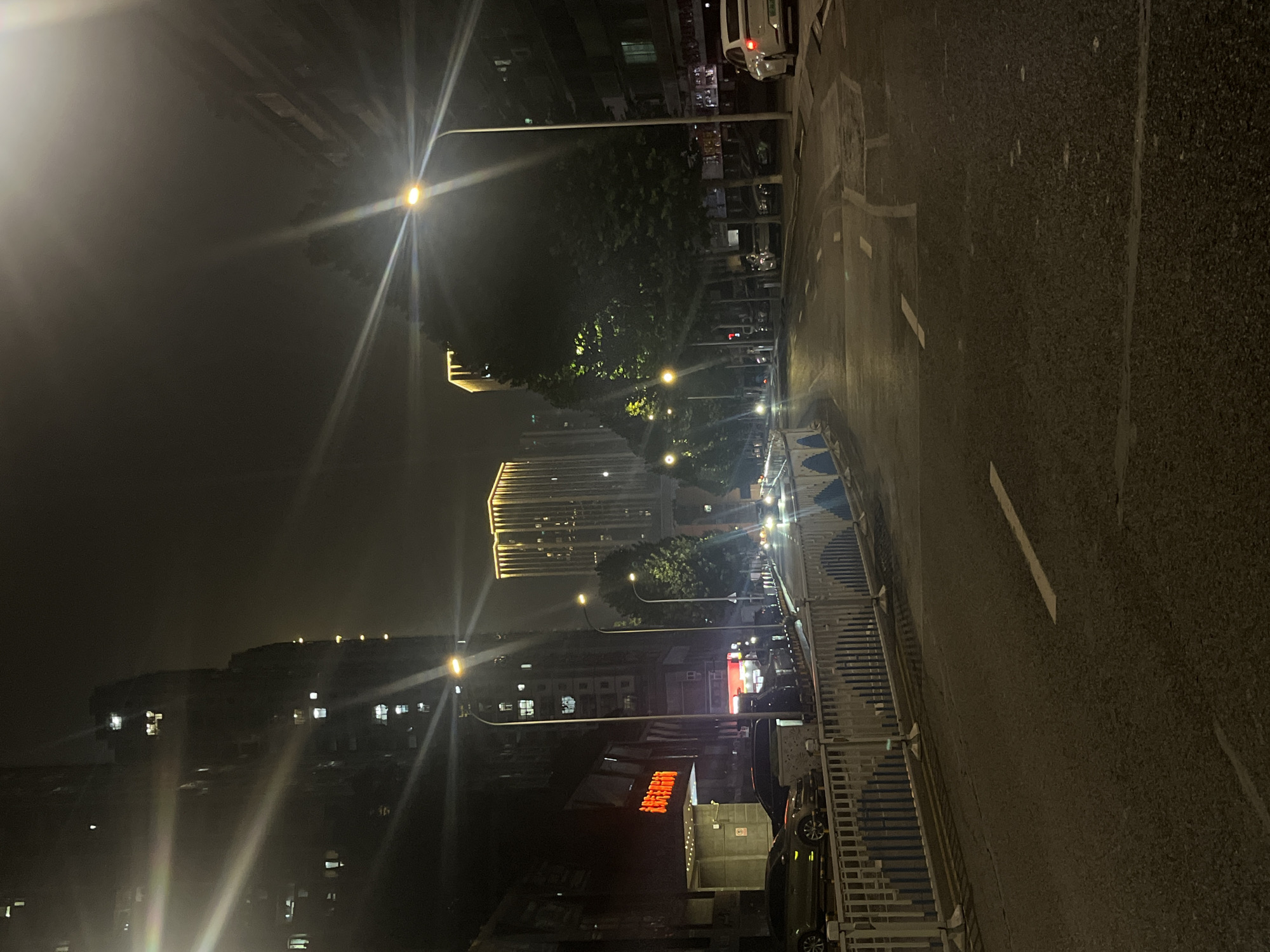 This is a nighttime photograph of a street in Yuzhong District, China. The street is lined with buildings and trees, and illuminated by streetlights and building lights. Several cars are parked along the side of the road. The road itself is wet, reflecting the lights. In the background, modern high-rise buildings with illuminated facades are visible. There are no people clearly visible in the image. The overall impression is of an urban environment at night, with a sense of stillness.