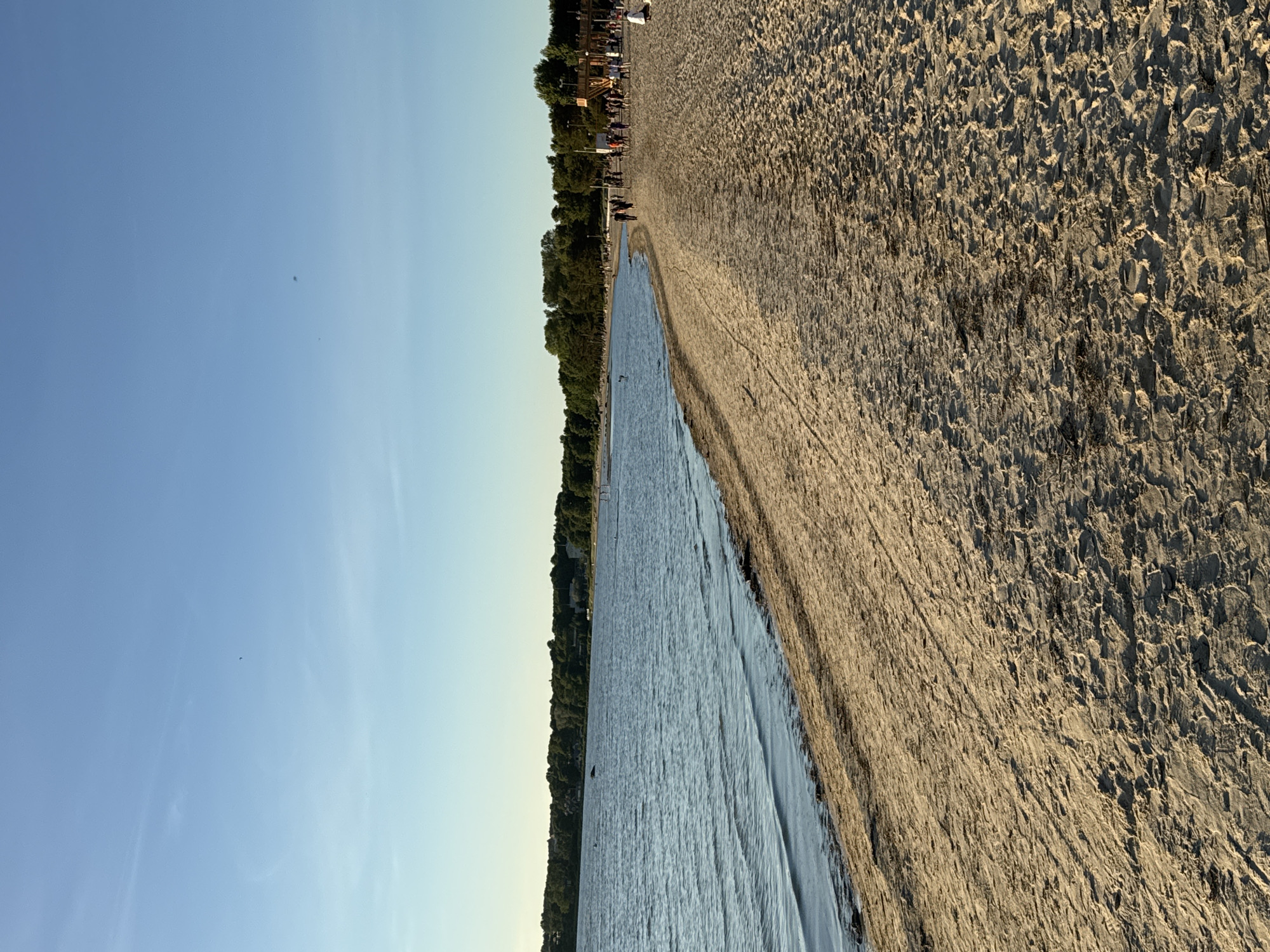 This is a photograph taken from a sandy beach looking out over a body of water towards a tree-lined shore. The sky is clear blue with a few wispy clouds. The beach is populated by several people walking along the shoreline and some individuals sitting or standing. The water appears calm with small ripples. In the distance, trees form a dense line along the coast, and there are some buildings visible amongst the trees. The sand is light brown and shows the texture of footprints and tire tracks. The lighting suggests it is either early morning or late afternoon, with the sun casting long shadows. The scene depicts a public beach area, likely a recreational spot within or near the city of Tallinn, Estonia, given the visual cues of a large body of water, significant tree coverage, and visible human activity.
