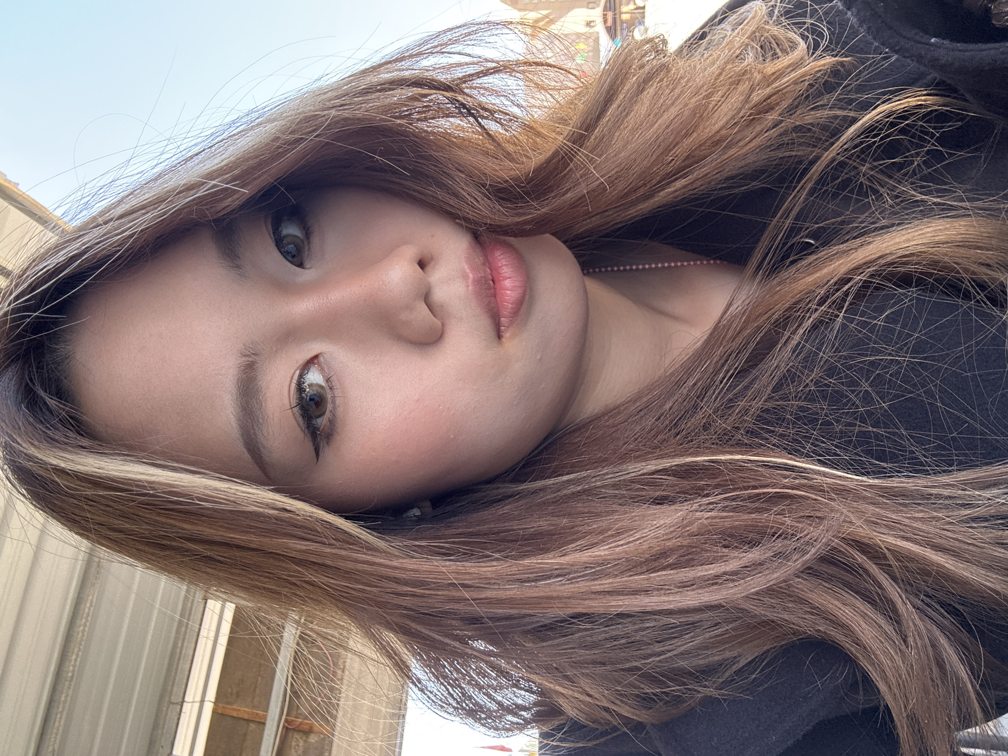 A close-up, low-angle portrait captures a young individual, likely female, with their head tilted significantly to the left, creating a sideways orientation. The subject has an oval face with prominent dark brown eyes enhanced by eyeliner and mascara, and well-groomed eyebrows. Her lips are slightly parted, revealing a light pink lipstick or gloss, and a subtle blush is visible on her cheeks.
Her long, wavy hair, a mix of reddish-brown with lighter, almost golden-blonde highlights, frames her face and flows across her shoulders. Some individual strands of hair are visible against the bright background. She is wearing a dark-colored top or jacket, and a delicate pink beaded necklace is visible around her neck.
The background is softly out of focus but suggests an outdoor setting under a clear, light blue sky, indicating daytime. Vague structural elements, possibly corrugated metal siding and distant buildings, are visible in the blurred background, hinting at an urban or semi-urban environment in Nantai, China. The subject's gaze is directed towards the viewer, conveying a neutral yet engaging expression. No visible text is present in the image.