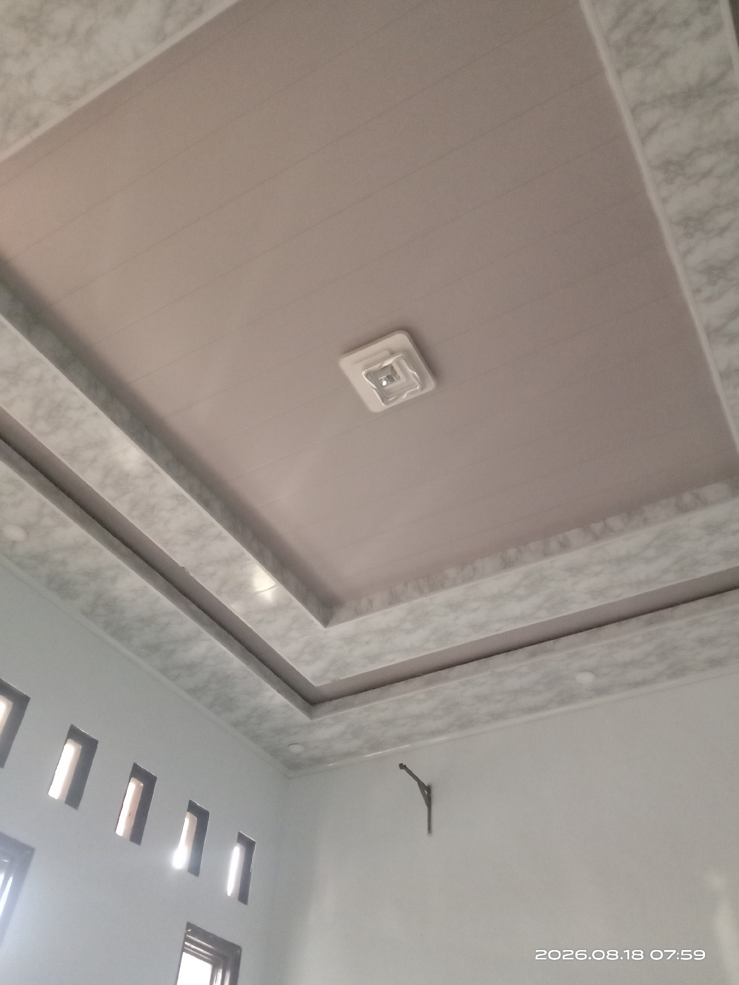 The image shows an interior view of a ceiling with a recessed lighting fixture. The ceiling is a light pink color with horizontal lines and has a decorative border with a marble-like pattern. Below the ceiling, a white wall is visible with several small rectangular openings that appear to be vents or decorative cutouts, allowing light to pass through. A small, dark metal bracket is attached to the wall. The timestamp "2026.08.18 07:59" is visible in the lower right corner of the image, suggesting a future date and time. There are no people or specific identifiable landmarks present in the image. The scene appears to be a residential or commercial interior.