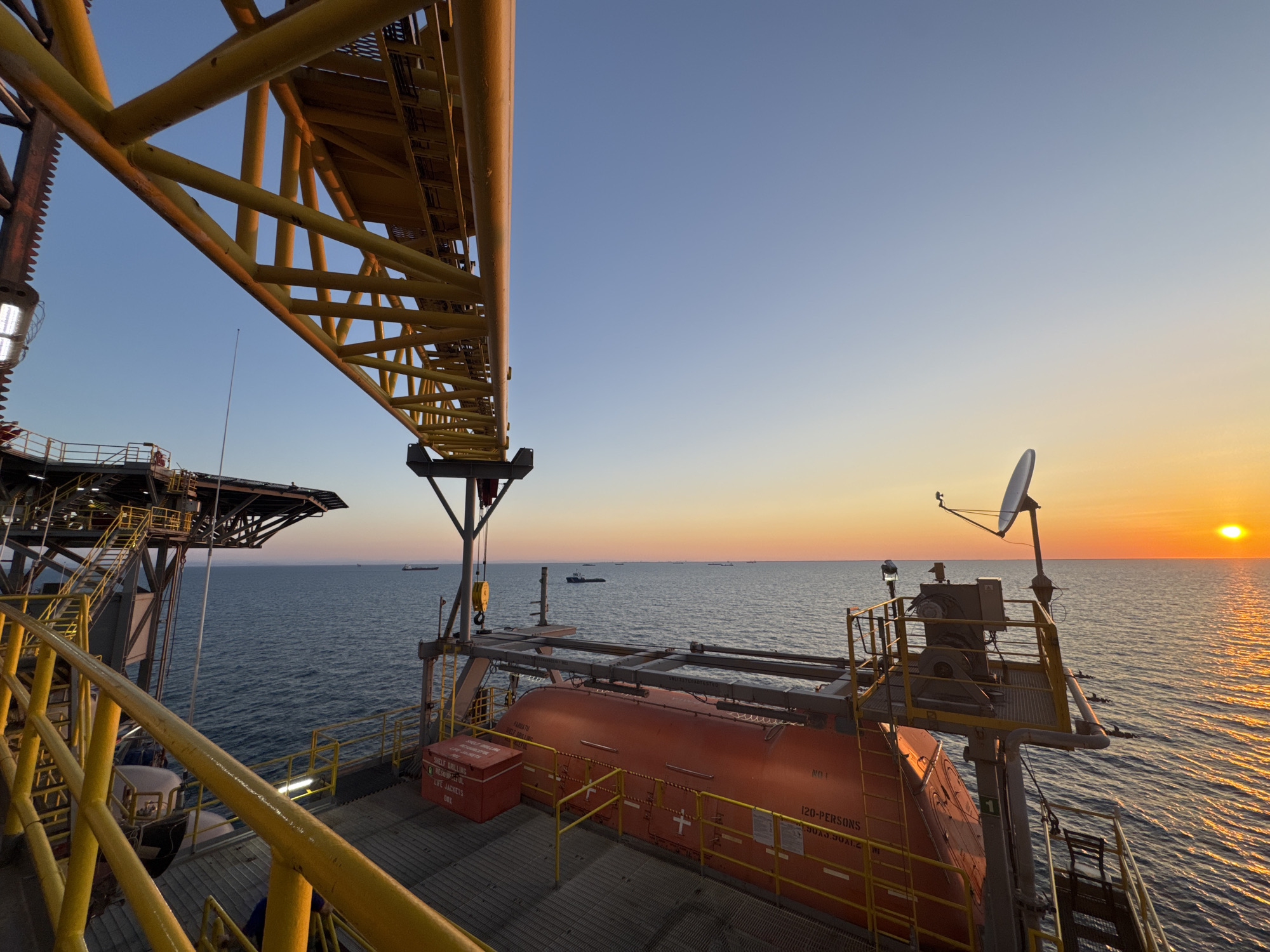 The image depicts an offshore oil rig at sunset. A large yellow structure, likely part of the rig's crane or framework, dominates the left side of the frame. Below this, a section of the rig's deck is visible, featuring yellow railings and a safety barrier. A bright orange lifeboat, labeled "120-PERSONS", is secured on the deck, along with a red storage container. The sea stretches out to the horizon, dotted with several ships in the distance. The sun is setting on the right, casting a warm orange glow across the water and sky. The overall environment is industrial and maritime, with the clear sky indicating fair weather. There are no people visible in the frame. The location is stated as Unknown, Italy, which implies an offshore setting as there is no indication of an urban landscape.