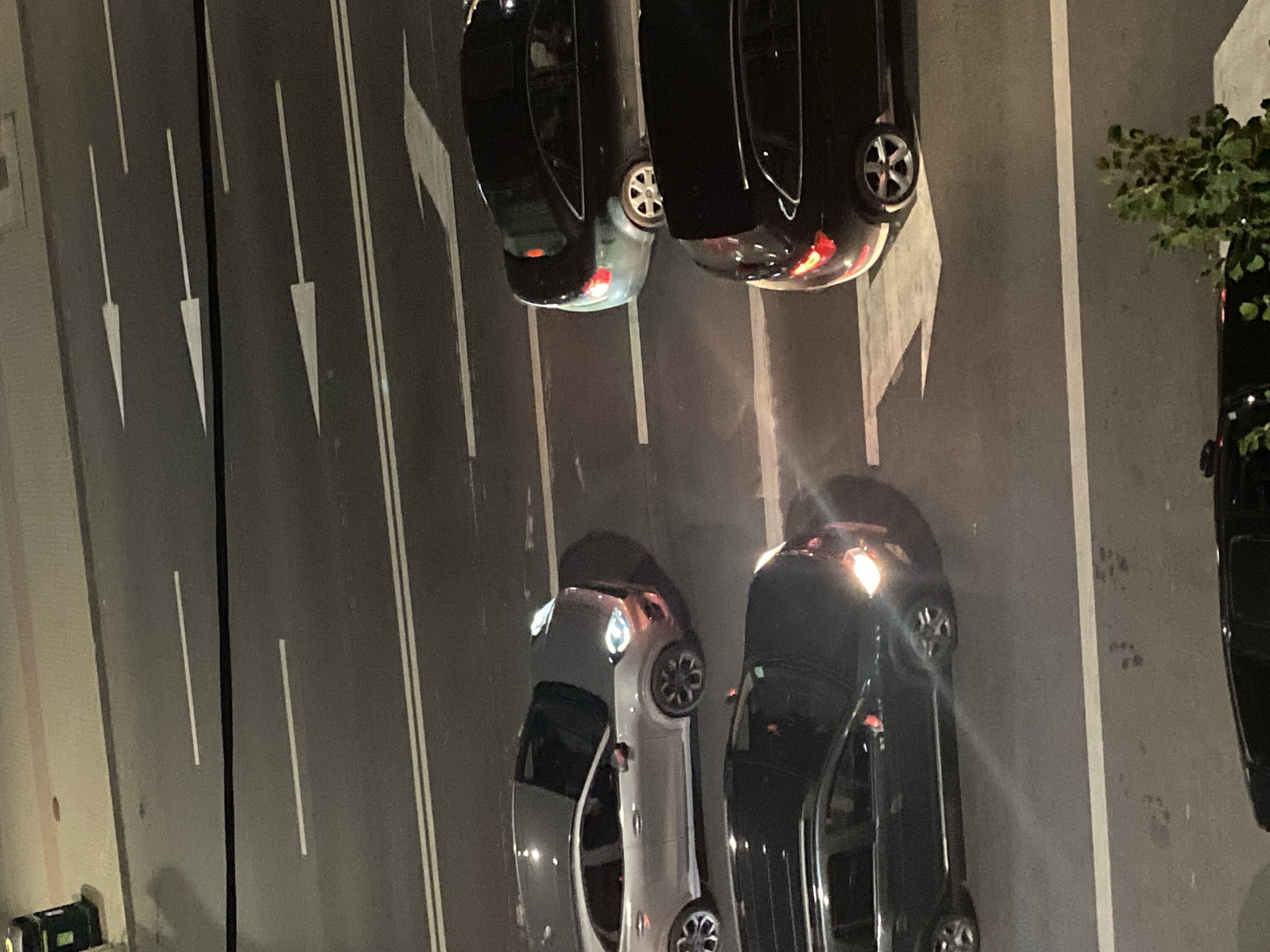 The image shows a nighttime aerial view of a road in Iași, Romania, likely from an elevated perspective such as a balcony or bridge. The road is paved and marked with white lane dividers and directional arrows. Four cars are visible on the road. From top to bottom, a silver SUV is parked in what appears to be a parking spot, facing left. To its right, a dark sedan is driving forward, its headlights off. Below the silver SUV and to the left, a black SUV is driving forward, its headlights on. To the right of the black SUV, another black SUV is driving forward, its brake lights illuminated. The environment is urban, with a sidewalk visible in the upper left corner featuring a dark green trash receptacle. The lighting suggests it is evening or night, with artificial illumination from streetlights and the vehicles themselves.