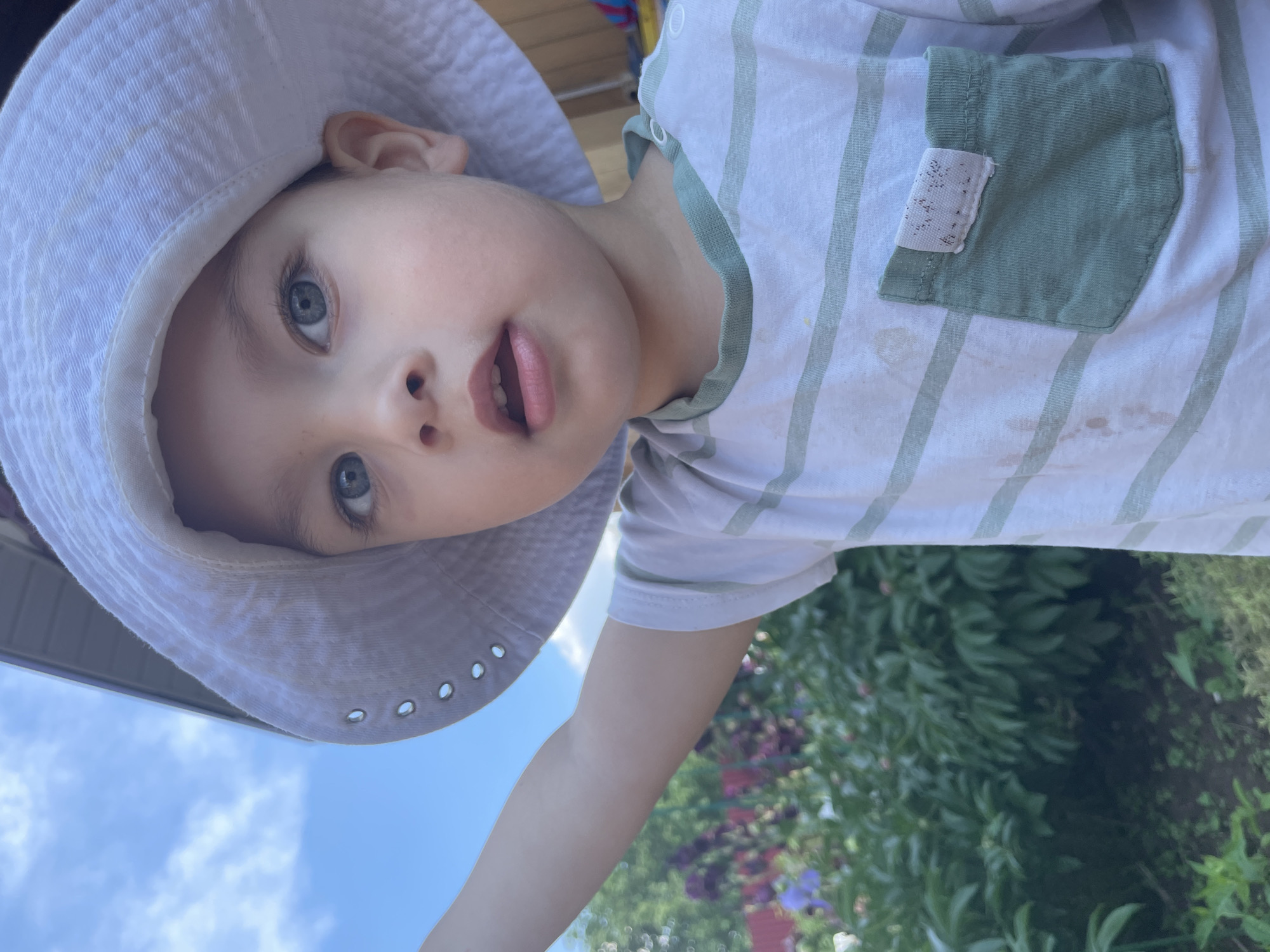 The image shows a close-up of a young child wearing a white bucket hat and a white t-shirt with light green stripes. The child has blue eyes and is looking towards the camera with their mouth slightly open. The child's left arm is extended, and their hand is out of frame. In the background, there is lush green foliage, possibly a garden, with some purple and pink flowers visible. The sky above is blue with scattered clouds. The setting appears to be outdoors, likely in a residential area, given the presence of a building overhang in the upper right corner. There are no visible elements to specifically place this image in Zav'yalovo, Russia, or any other specific city.