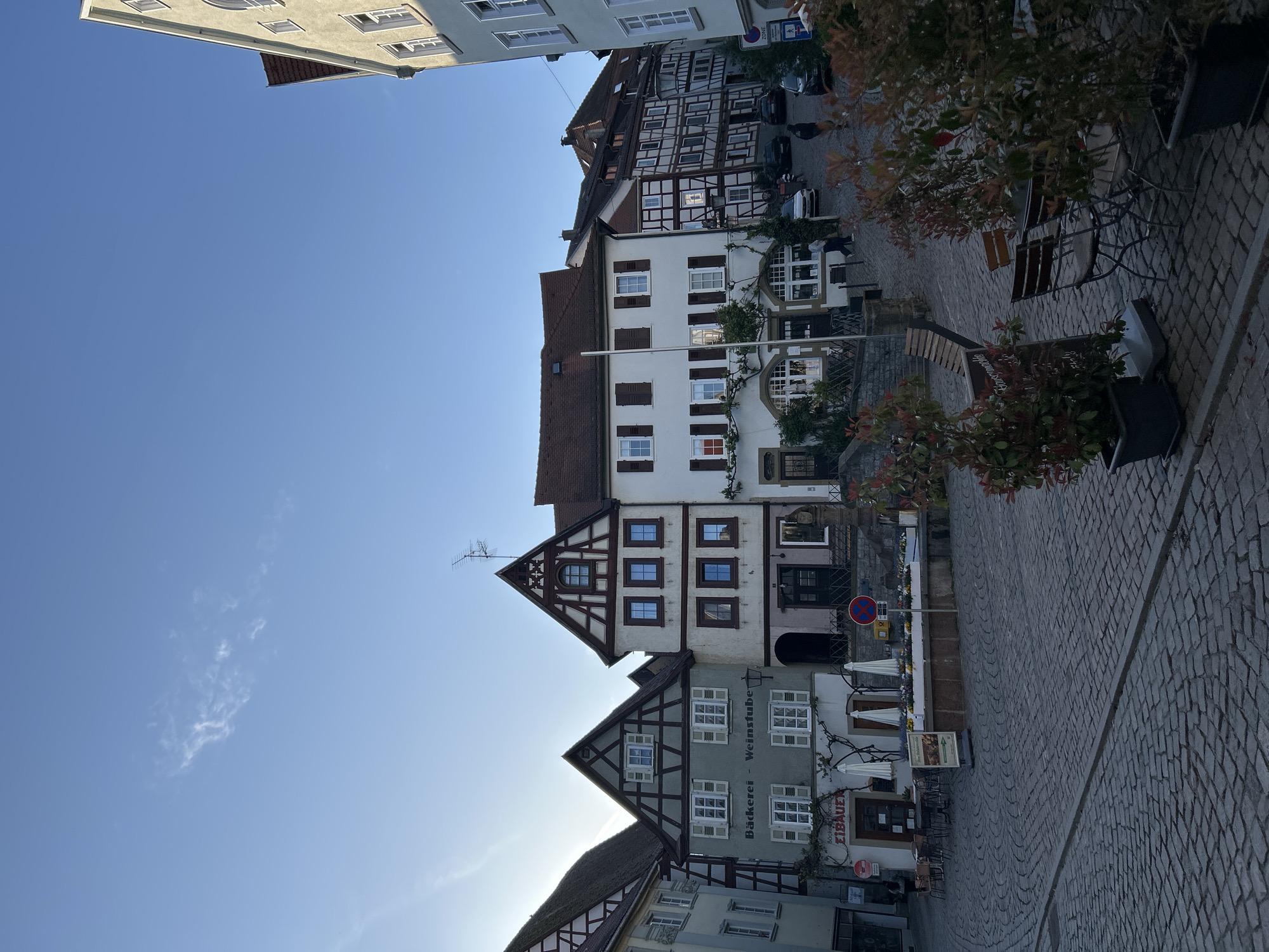 The image depicts a street scene in an unidentified city. Several buildings with traditional European architecture, characterized by timber framing and pitched roofs, line the street. One building displays signage in German that reads "Bäckerei - Weinstube" and "Eibauer". The street is paved with cobblestones. Outdoor seating, including chairs and tables, is visible. The sky is clear and blue, suggesting a sunny day. No people are directly visible in the frame.