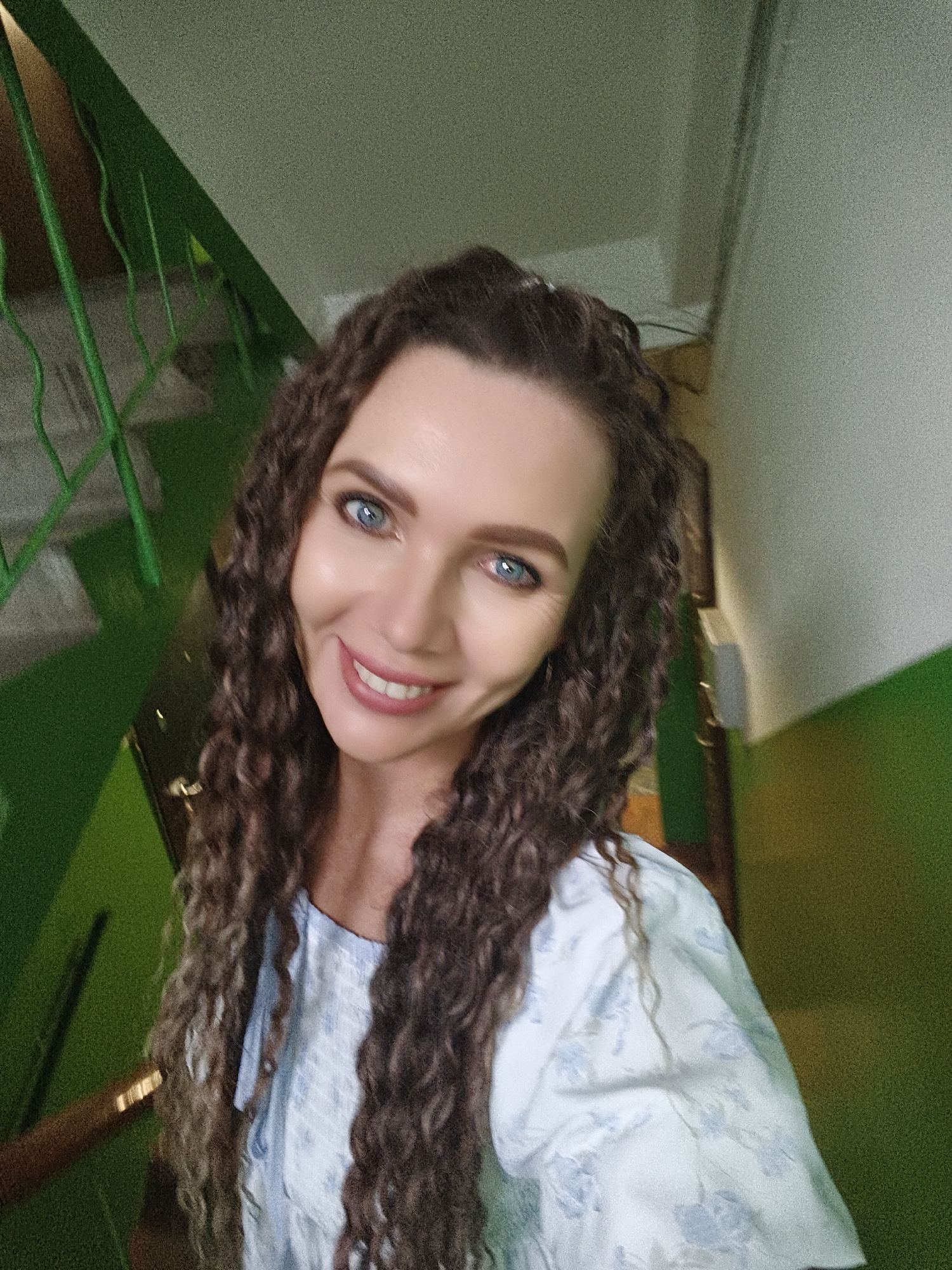 A selfie of a woman with long, curly brown hair and blue eyes is taken from a slightly elevated perspective. She is smiling and looking at the camera. She is wearing a light-colored, floral-patterned top. Behind her, a green metal railing of a staircase is visible on the left, with green walls and a beige wall to the right. The lighting suggests an indoor environment, possibly a stairwell in an apartment building in the city of Chaikovsky, Russia.
