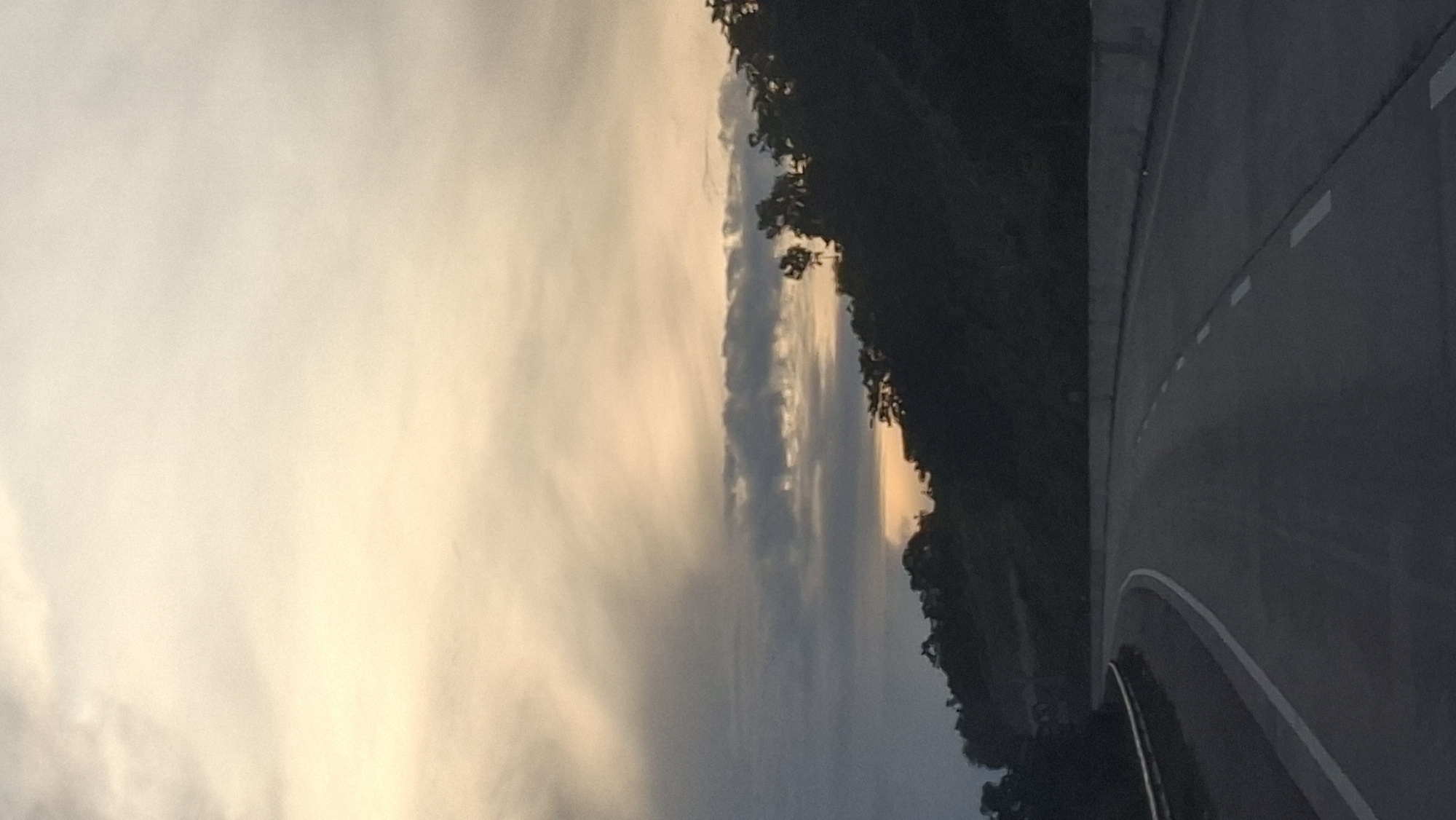 The image depicts a road curving to the left, bordered by a concrete barrier on the right and a metal guardrail on the left. The road is paved and has white lane markings. On the right side of the road, a dense hillside covered in dark foliage rises sharply, silhouetted against a dramatic sky. The sky is filled with a gradient of colors, transitioning from a pale yellow and cream at the top to darker grays and blues closer to the horizon, with streaks of clouds. A sliver of orange light, suggesting a sunset or sunrise, is visible peeking through the clouds and above the treeline. The overall impression is that of a highway or major road passing through a natural, hilly landscape, likely at dusk or dawn. The location is identified as Lamtamot, Indonesia.
