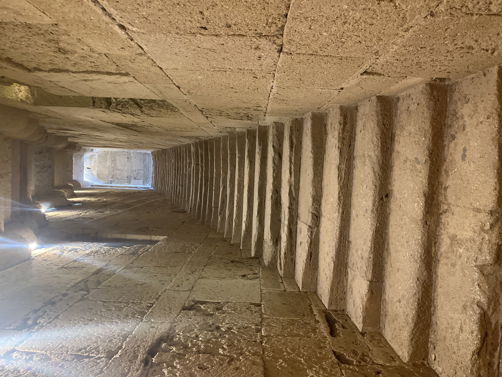 The image depicts a stone staircase ascending within an ancient structure, likely part of the Cathédrale Saint-Pierre-et-Saint-Paul in Villeneuve-lès-Maguelone, France. The walls, constructed from large, rough-hewn stone blocks, are ancient and weathered, showing signs of age and erosion. Sunlight streams from the top of the staircase, creating dramatic shadows and highlighting the texture of the stone. The steps themselves are broad and made of the same heavy stone, worn smooth by time and possibly foot traffic. The overall atmosphere is one of historical significance and rugged construction. There are no people or immediate signs of activity in the frame, suggesting a quiet, preserved historical site. The perspective draws the viewer's eye upwards, following the path of the stairs into the light.