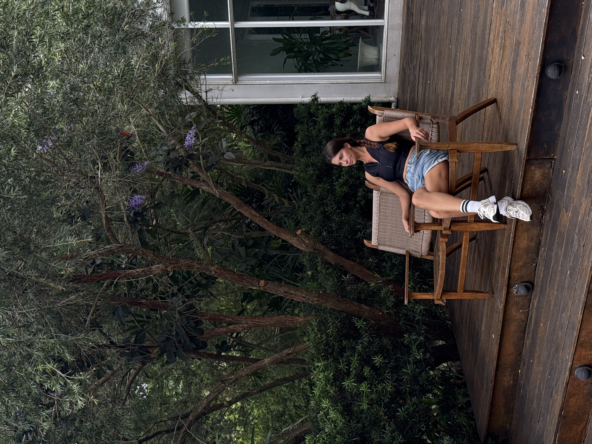 A woman is sitting on a wooden deck outside a house. She is wearing a black sleeveless top, denim shorts, white socks, and athletic shoes. She is sitting in a wicker chair with a wooden frame. The deck is made of dark wood planks. Behind the woman, there are lush green bushes and trees. A large window with a view into the house is visible to her right. The overall impression is one of a peaceful, natural setting. The location is identified as Ko Samui District, Thailand.