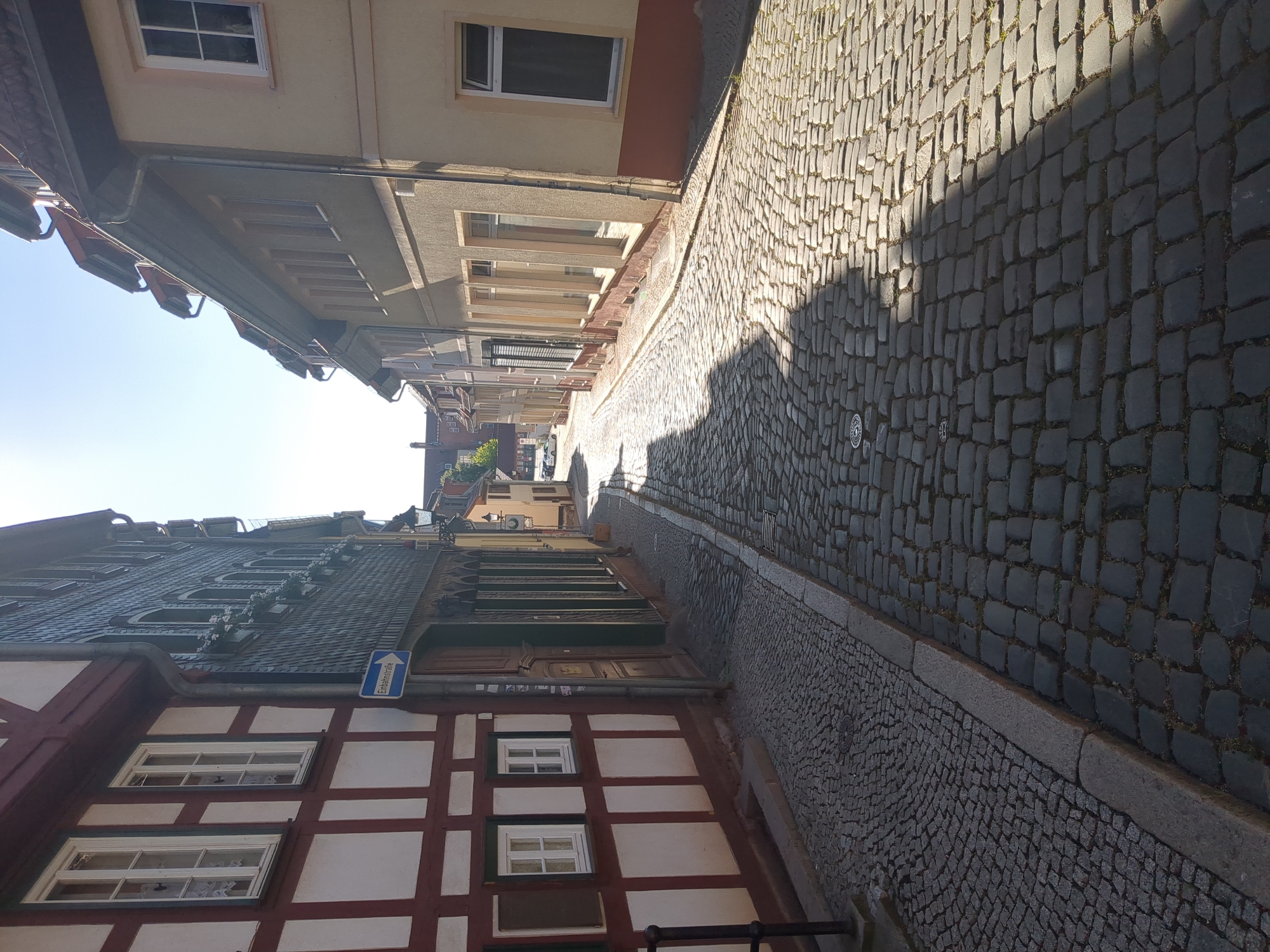 The image shows a narrow cobblestone street in Nordhausen, Germany. The street is lined with historic buildings, primarily on the left and right sides, with a gentle curve leading into the distance. The building on the left features traditional half-timbered architecture with white infill and dark red timber framing, along with dark green shutters on some windows. A blue street sign with white text and an arrow pointing to the right is visible on this building, indicating a one-way street. The building on the right has a lighter, stuccoed facade. The cobblestone pavement is worn, suggesting age and frequent use. Sunlight casts distinct shadows across the street, indicating a bright, sunny day. The sky is clear and blue. No people are visible in the immediate scene. The overall impression is of a well-preserved historical street within a German town.