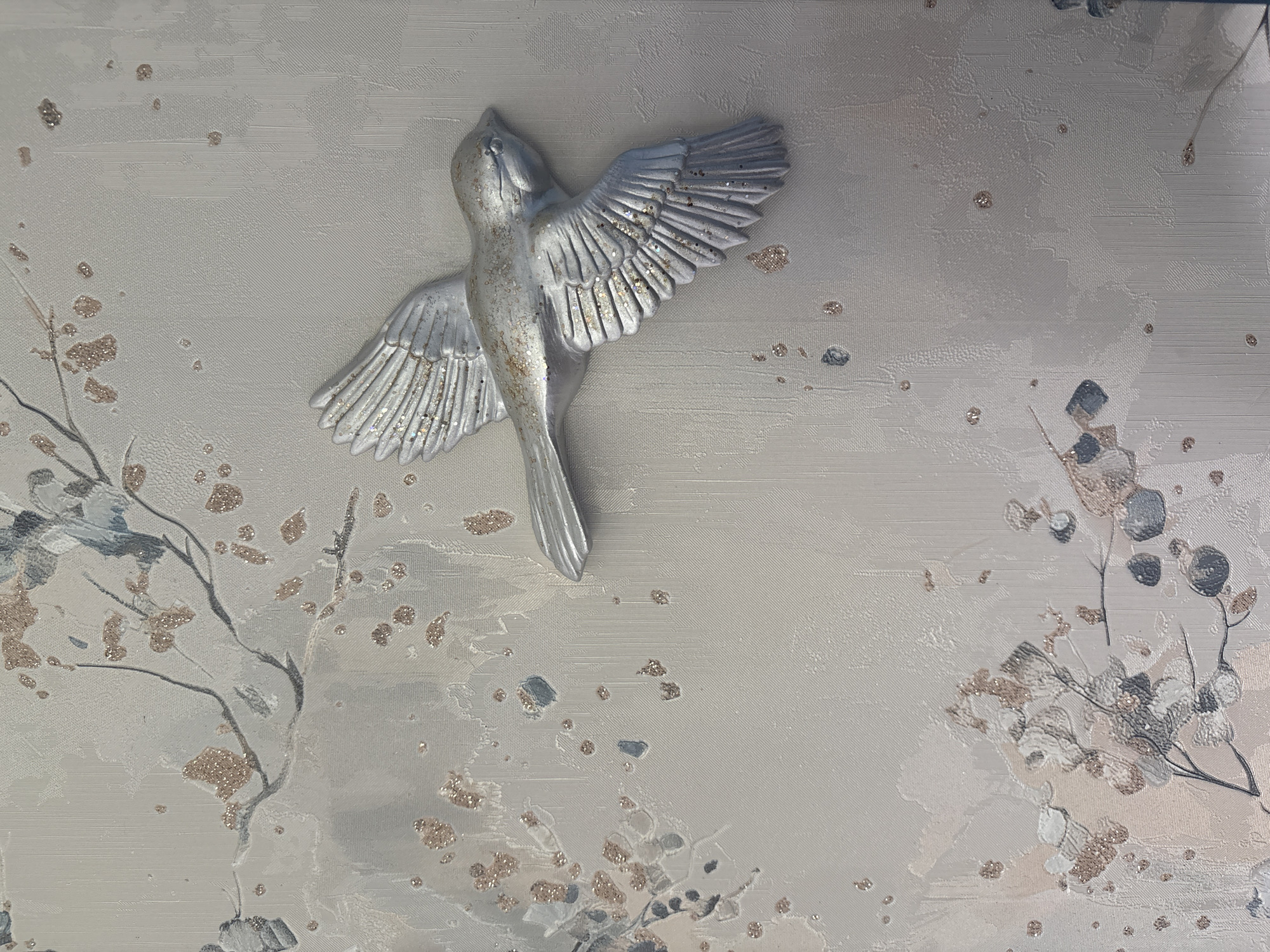 The image displays a textured wallpaper with a delicate floral and branch pattern, accented with flecks of glitter. A three-dimensional silver bird sculpture, also subtly shimmering with glitter, is affixed to the wallpaper, appearing to be in mid-flight with its wings outstretched. The overall aesthetic is soft and elegant, with a color palette dominated by muted grays, whites, and pale pinks. The scene does not provide any information about a specific location within the city of Дольче Вита Кп, Russia, as it appears to be an indoor decorative element. There are no people or actions depicted.